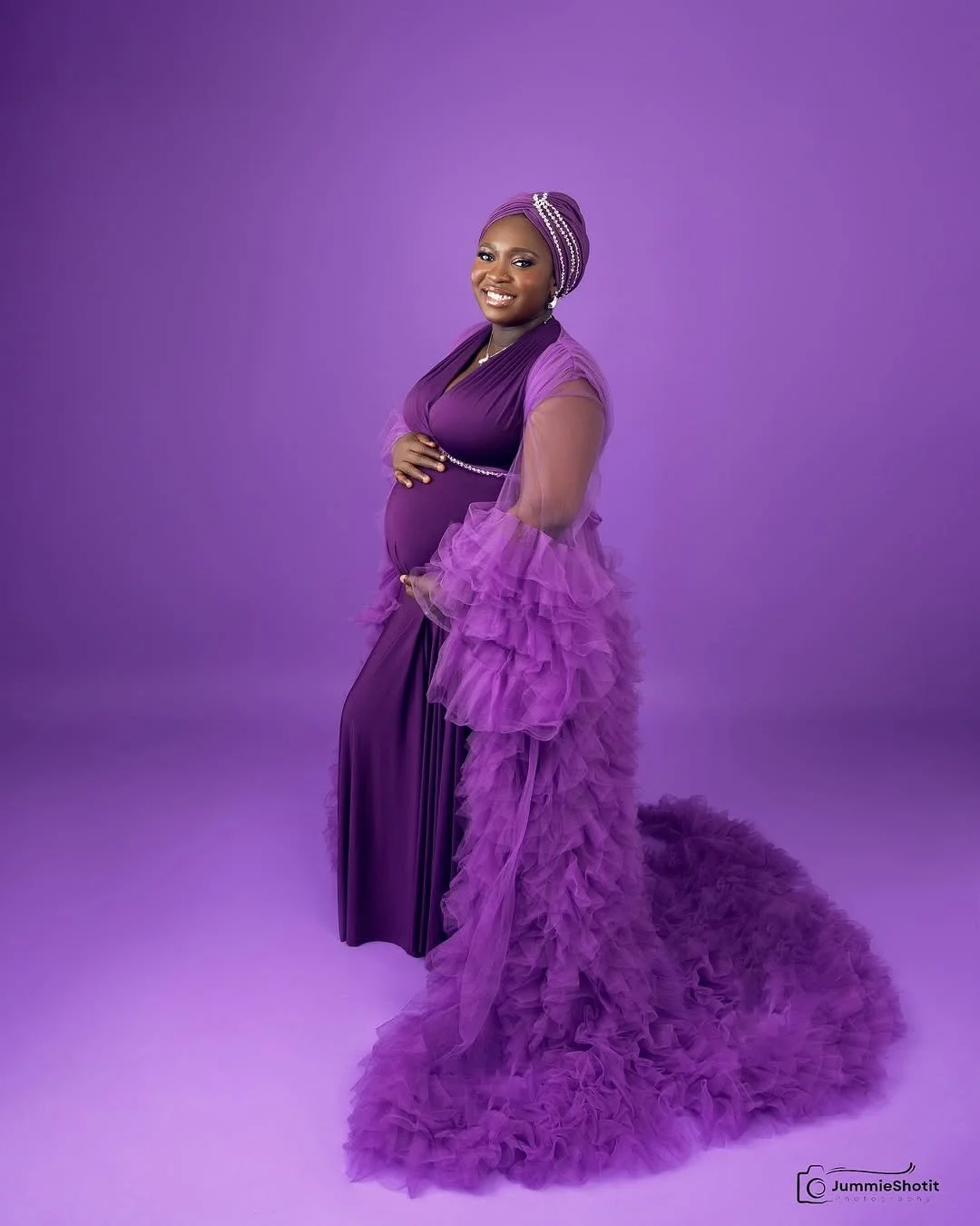 A pregnant woman wearing a silky dark purple dress and fluffy tulle robe in studio maternity photoshoot