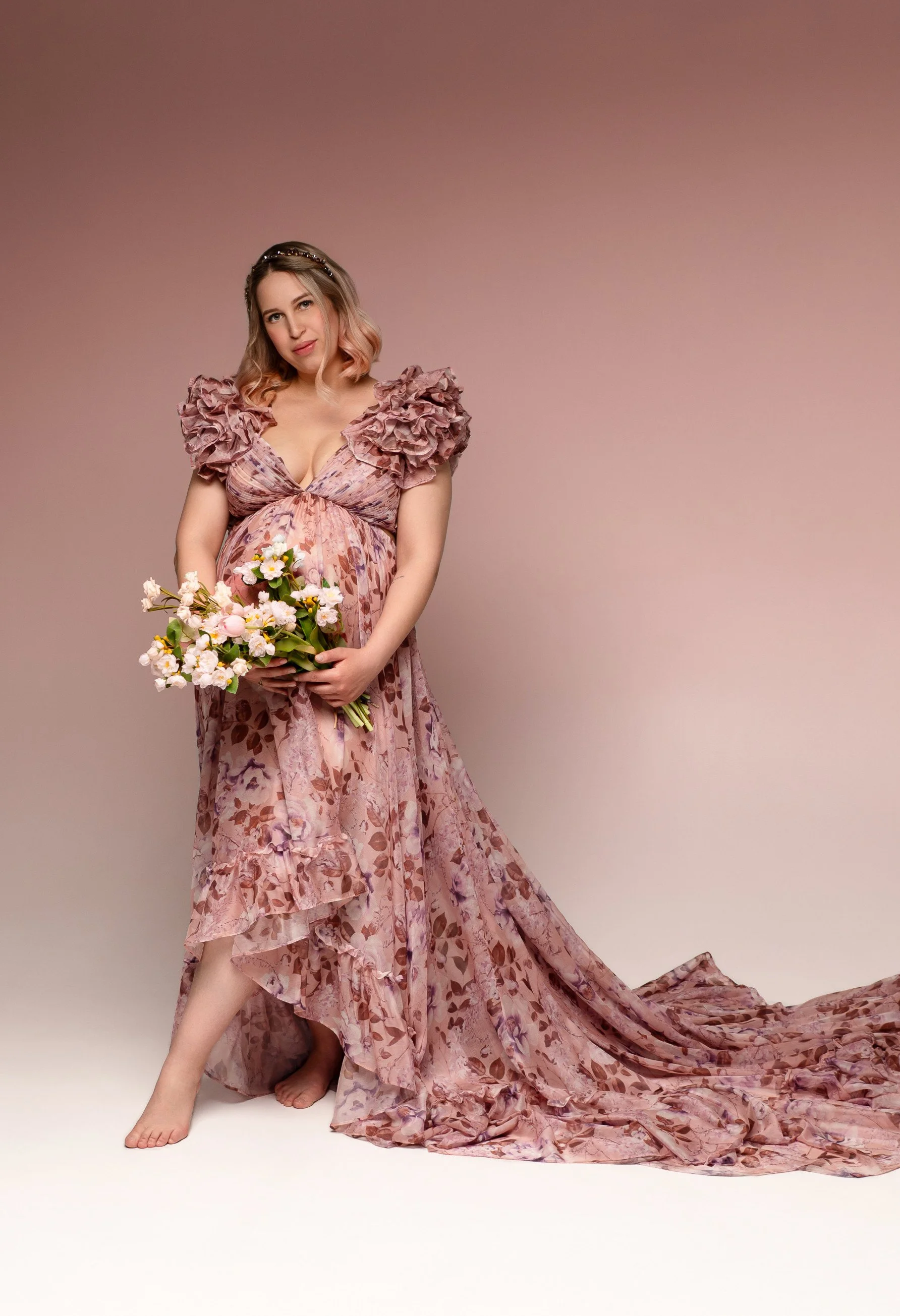 A pregnant woman wearing a pink floral organza gown with off-the-shoulder v-neck corset, holding flowers in a maternity photosession in Calgary