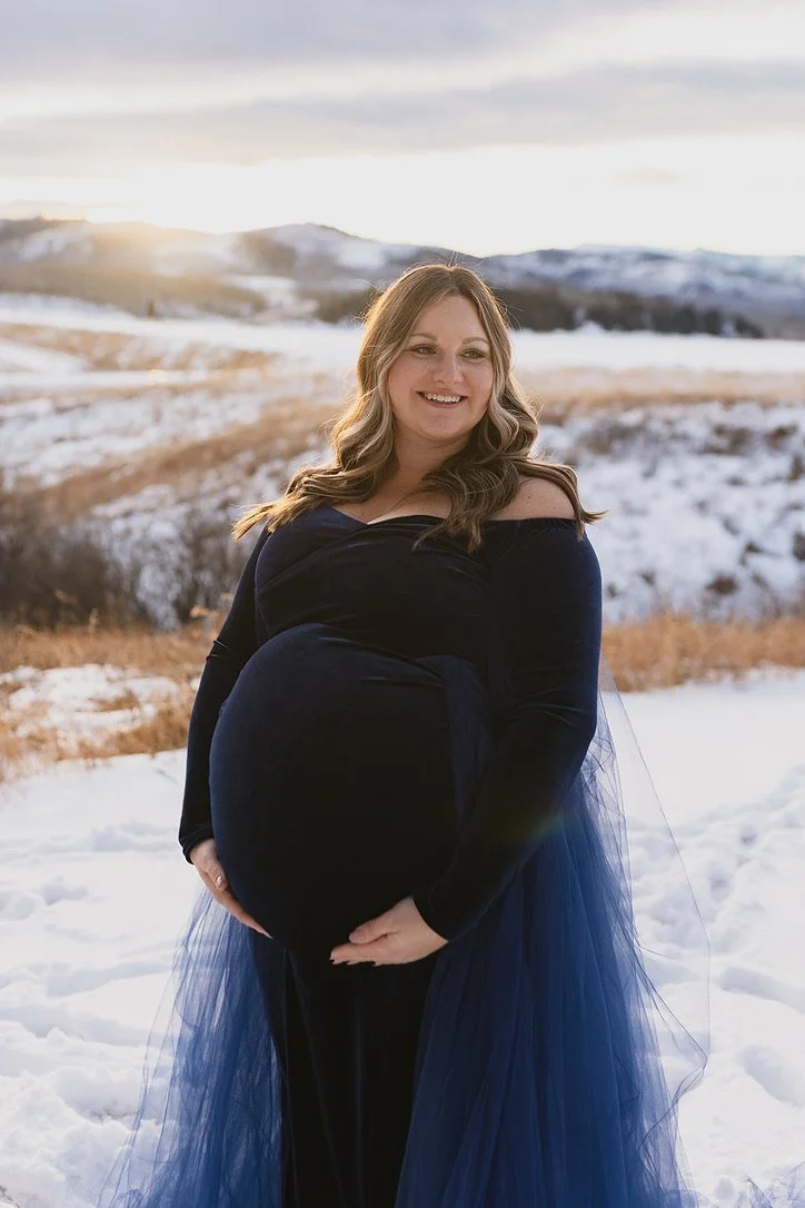 A pregnant woman wearing a navy dress and blue tulle overskirt in outdoor Calgary maternity photoshoot