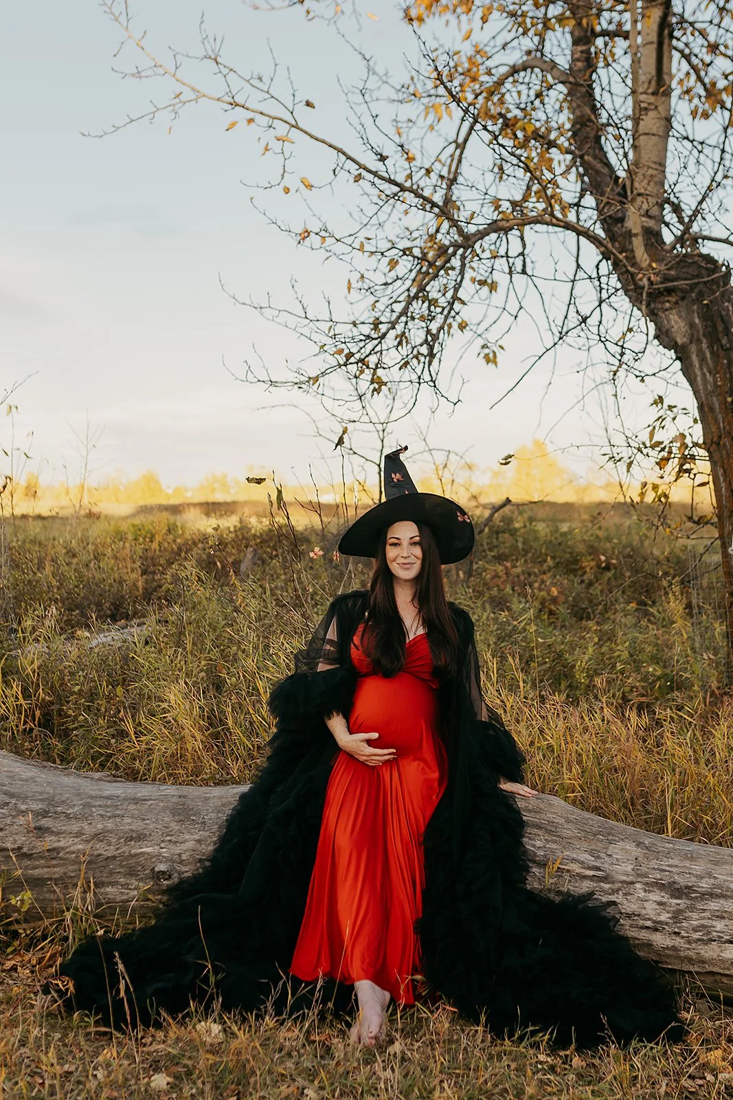 A pregnant woman wearing an orange infinity dress with a black tulle robe and witch hat in outdoor autumn maternity photosession