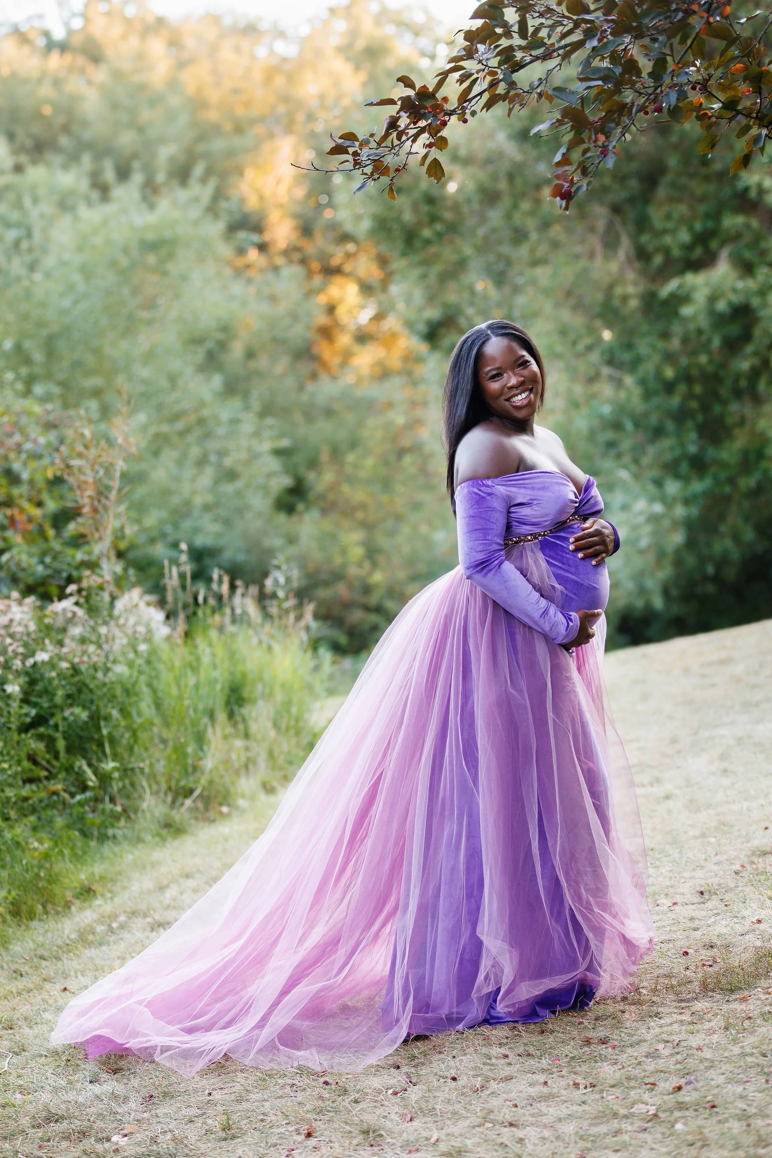 A pregnant woman in an outdoor Calgary maternity photoshoot, wearing a violet velvet dress with a floor length skirt