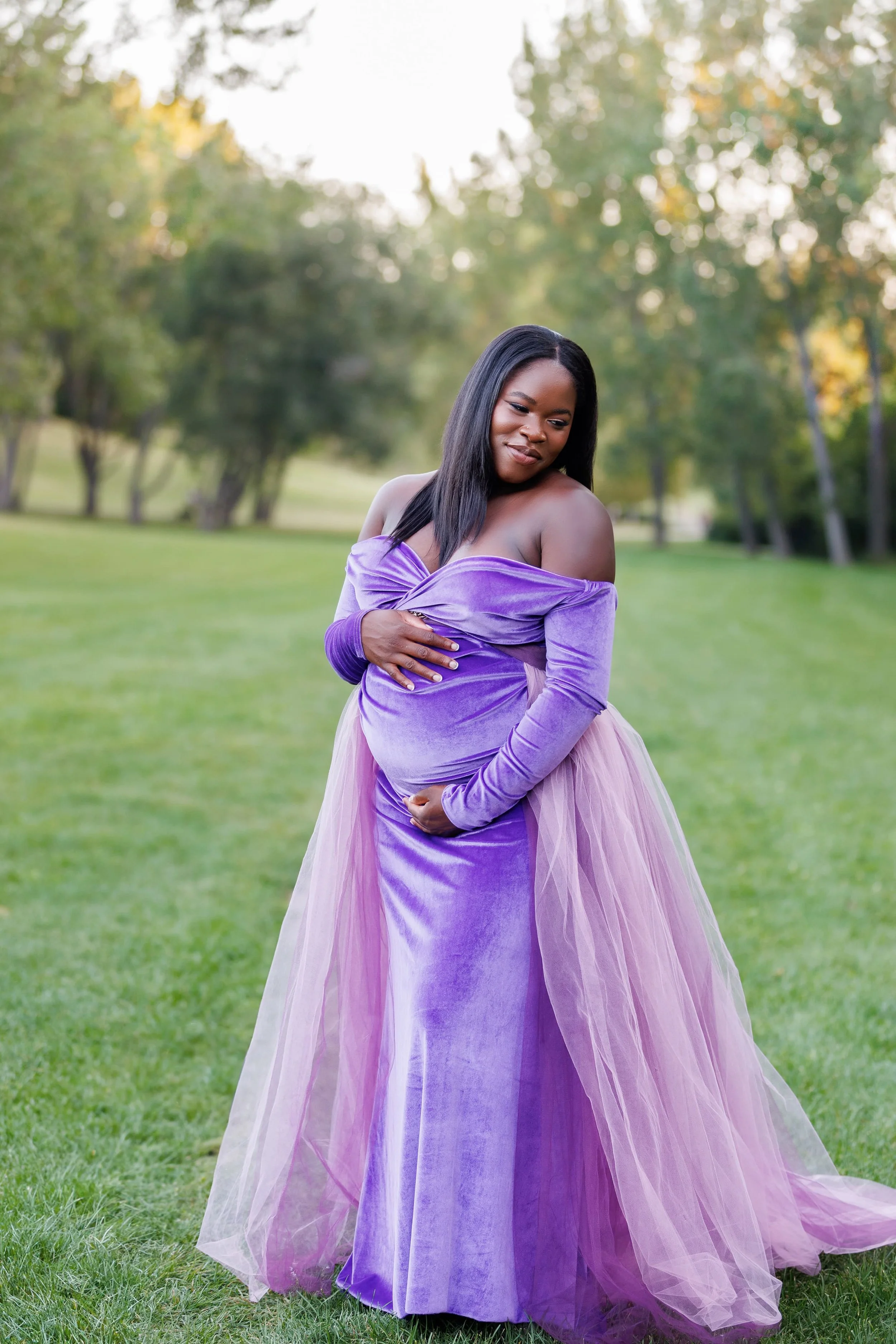 A pregnant woman in an outdoor Calgary maternity photoshoot, wearing a violet velvet dress with a floor length skirt