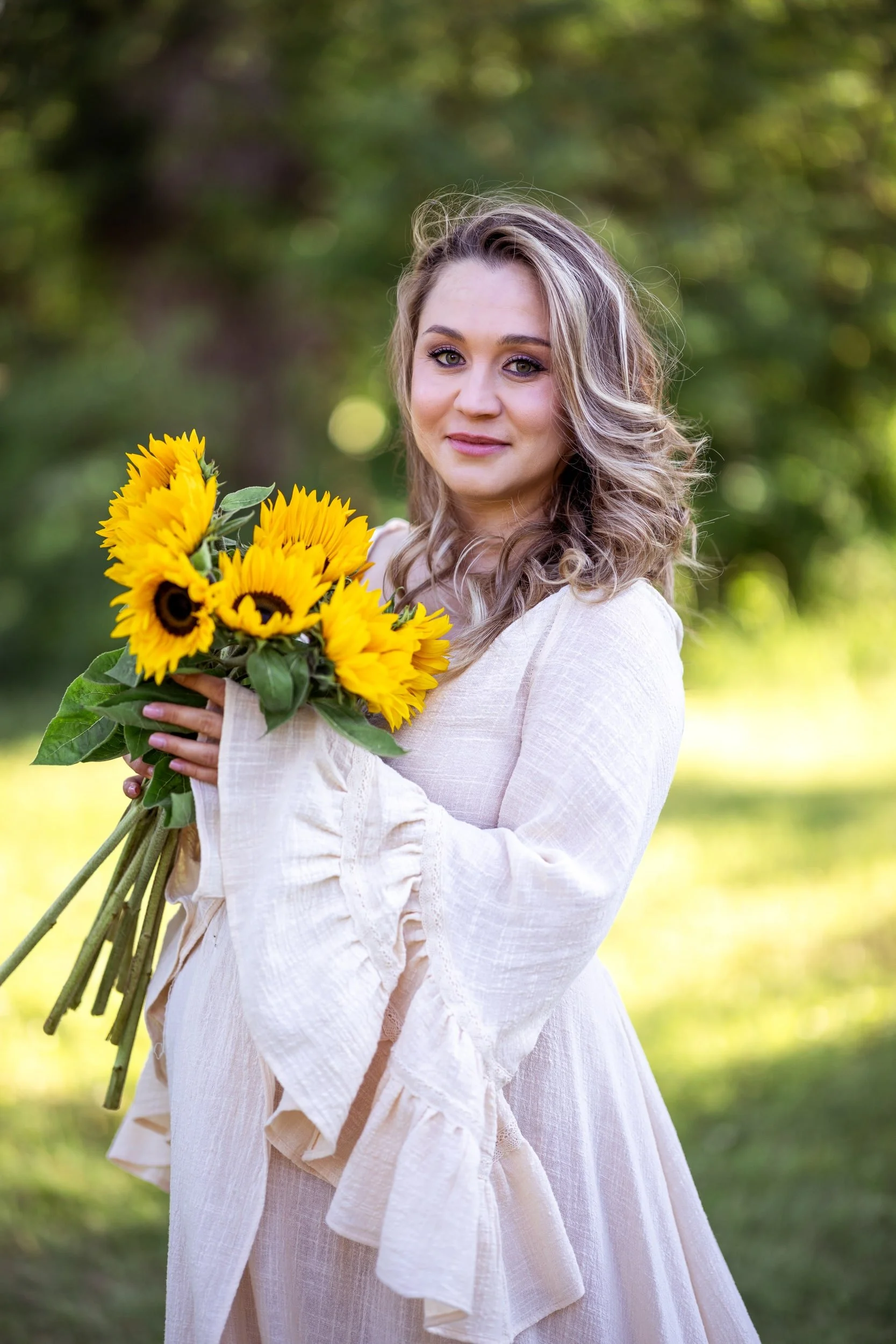 Portrait of pregnant woman with sunflowers, wearing a cream white boho gown with long ruffled sleeves and v-neckline, in outdoor summer photoshoot in Calgary