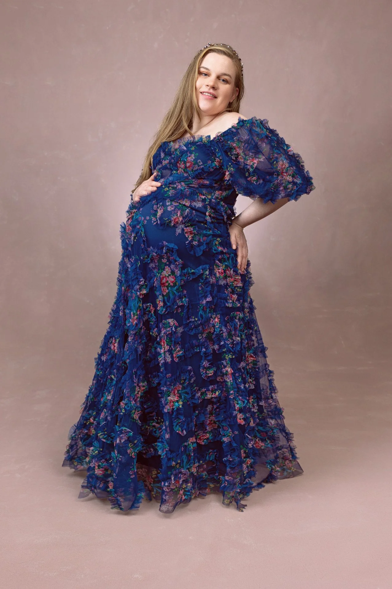 Pregnant woman wearing a navy blue dress with floral patterns and a lot of tulle texture, a maxi mermaid skirt, and off-the-shoulder sheer sleeves