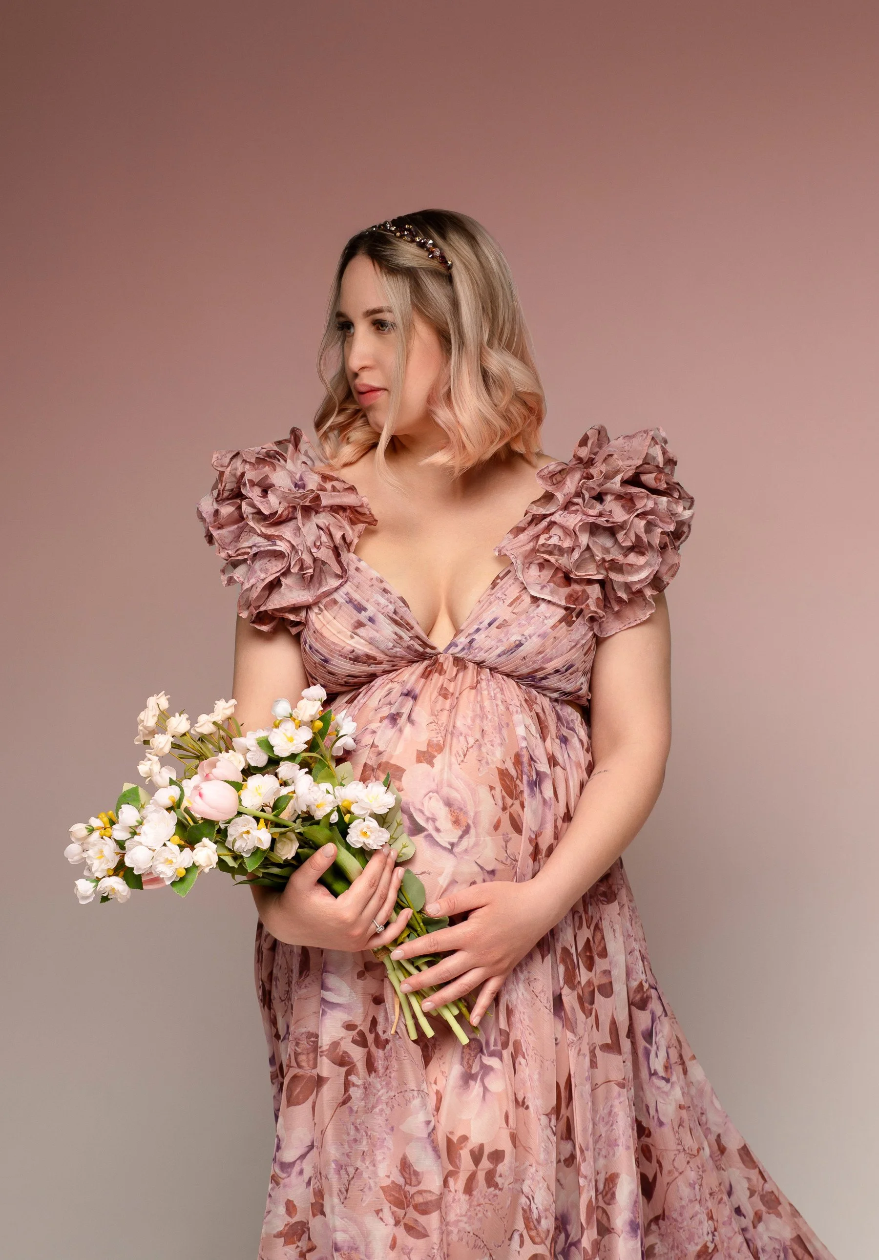 Portrait of a pregnant woman wearing a pink floral organza gown with off-the-shoulder v-neck corset, holding flowers in a maternity photosession in Calgary
