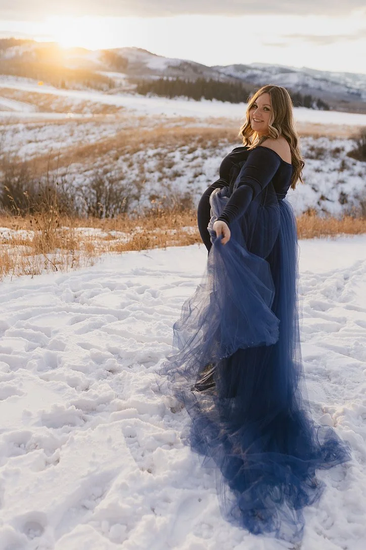 A pregnant woman wearing a navy dress and blue tulle overskirt in outdoor Calgary maternity photoshoot