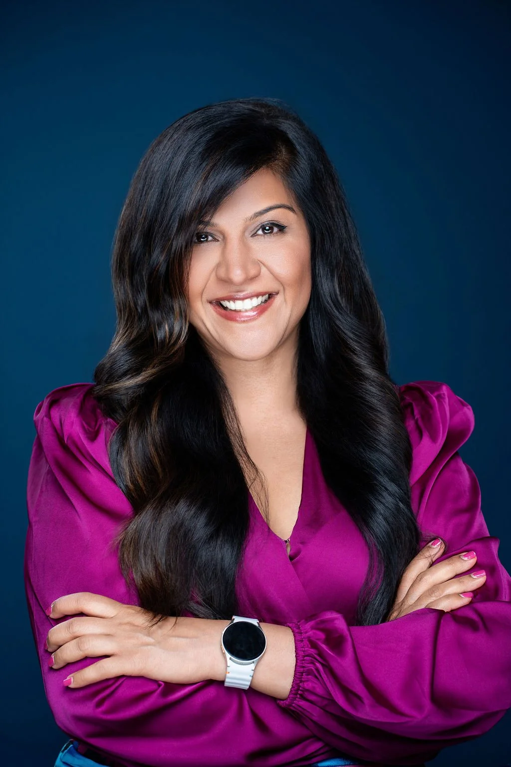 A woman with long dark hair smiling, wearing a dark blue blazer over a white top, standing against a light gray background.