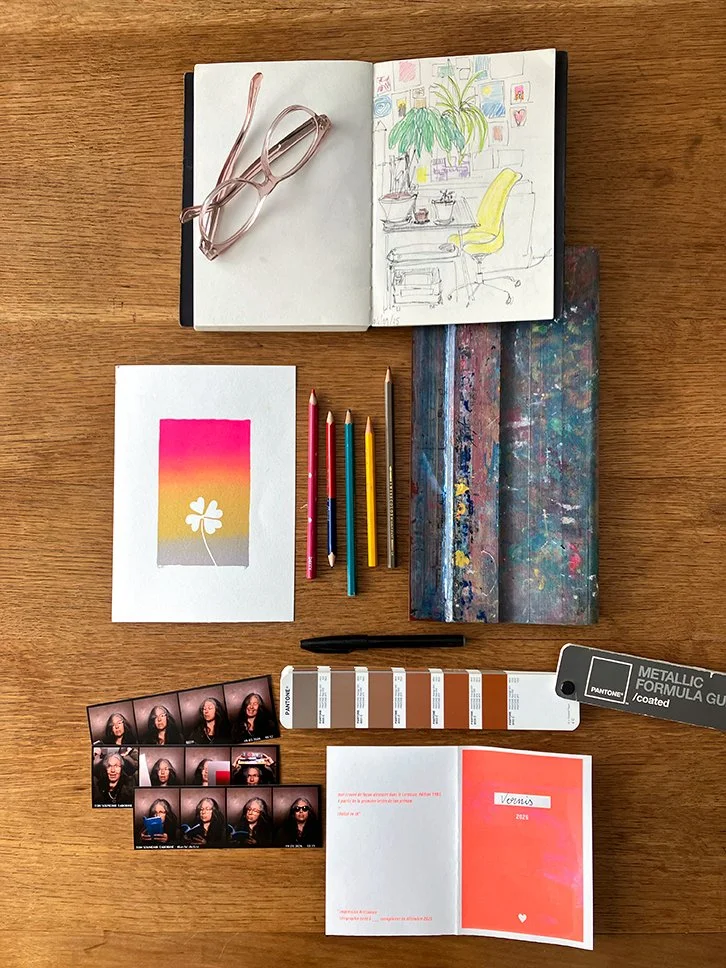 Une table en bois avec des objets d'art et de bureau, y compris un carnet de croquis ouvert avec un dessin d'un bureau avec une plante et un fauteuil, des crayons de couleur, une palette de peinture, des photos de portrait, un échantillon de couleurs Pantone, et un carnet avec une inscription 'Vœux 202'.