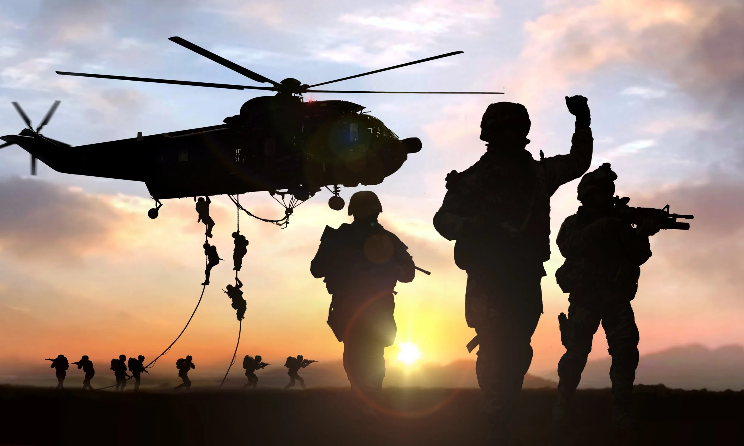 Silhouette of soldiers disembarking from a helicopter using ropes at sunset, with armed soldiers on the ground.