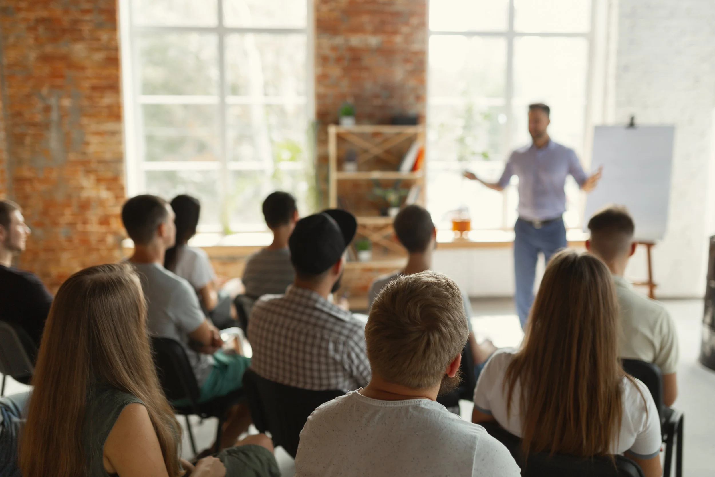 A group of people seated in a room, attentively listening to a speaker standing near a paper easel. The room has exposed brick walls and large windows.