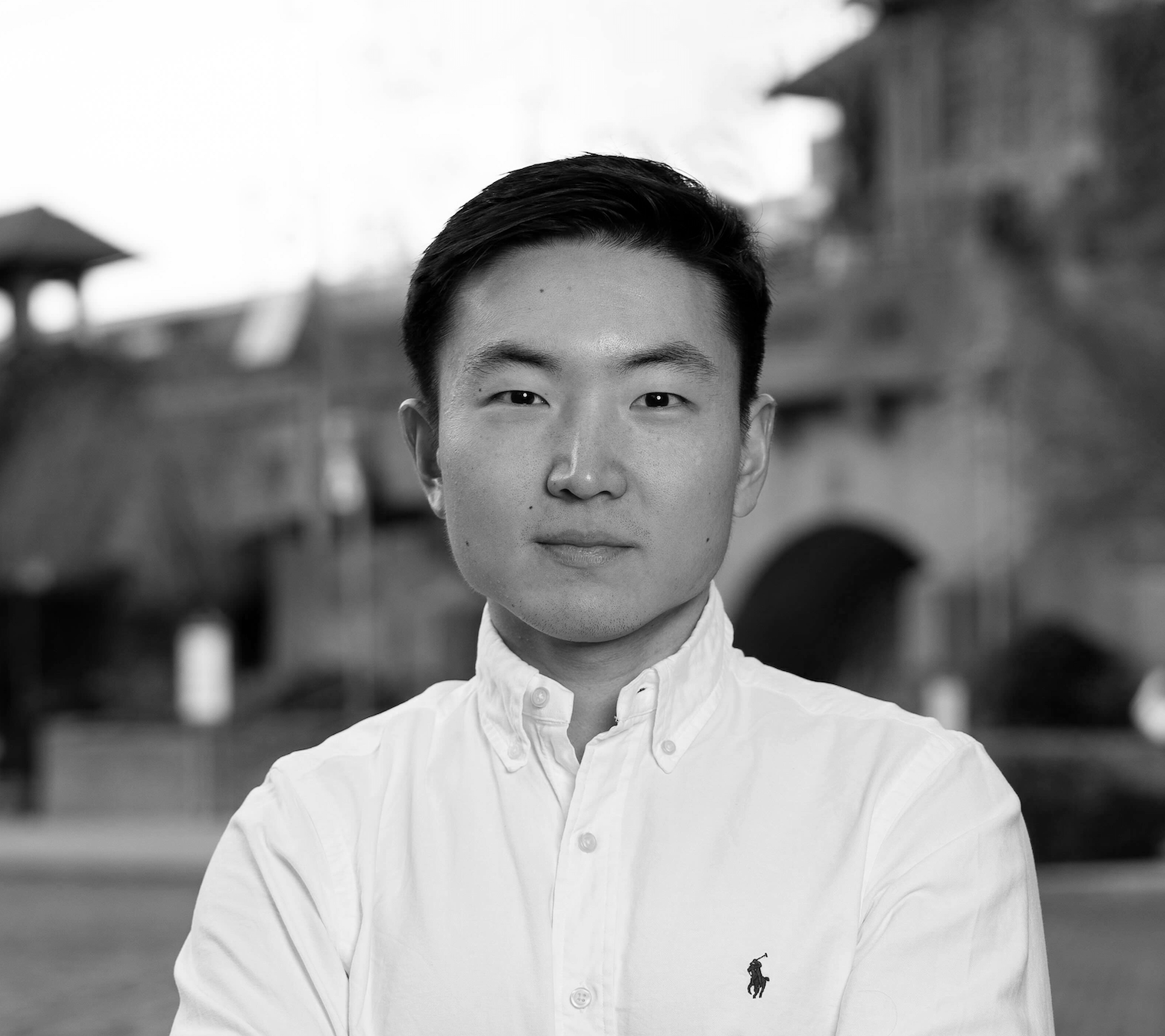 A black and white portrait of a young man with short dark hair, wearing a white button-down shirt with a small logo on the left chest, standing outdoors with a blurred background of a brick bridge and buildings.