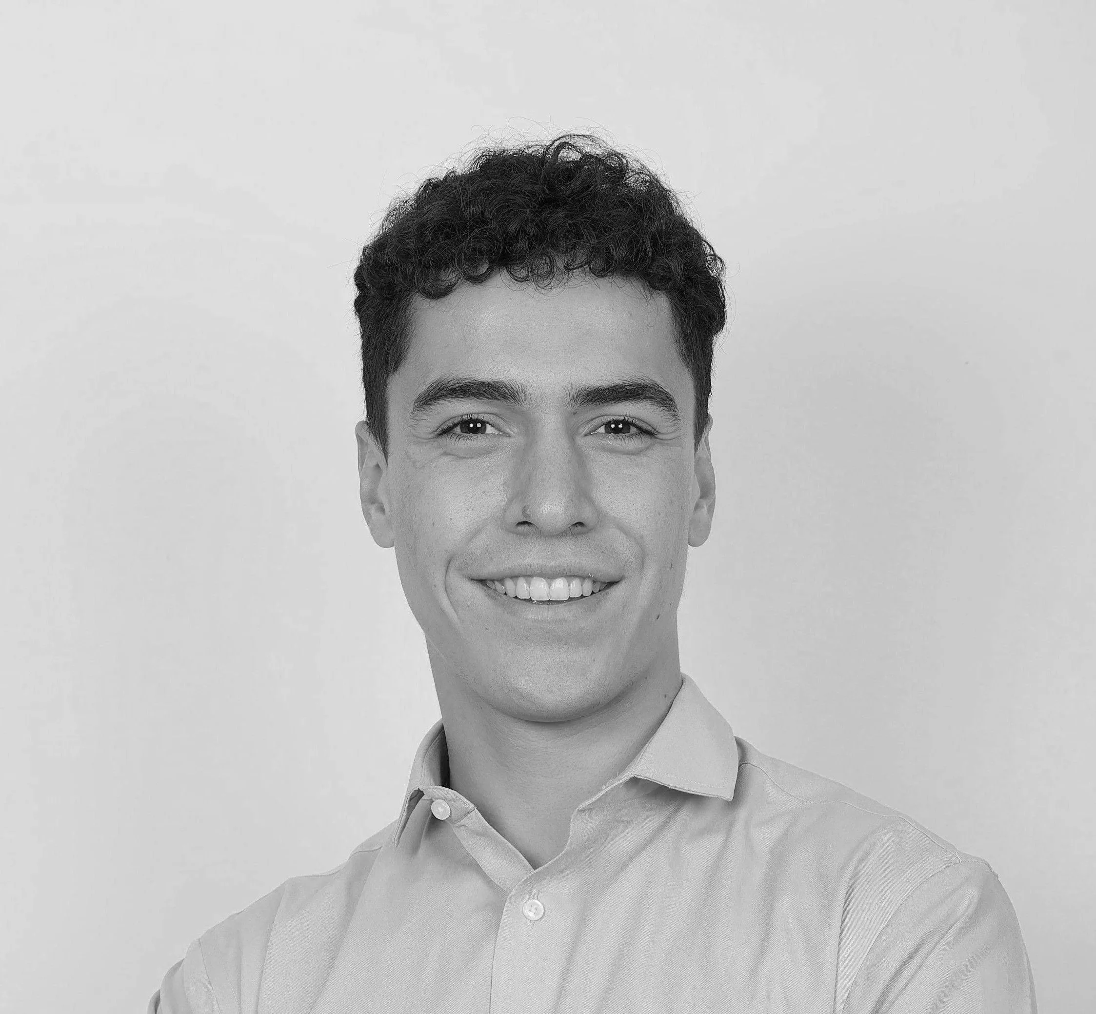 A black-and-white photo of a smiling young man with curly hair, wearing a collared shirt, against a plain background.