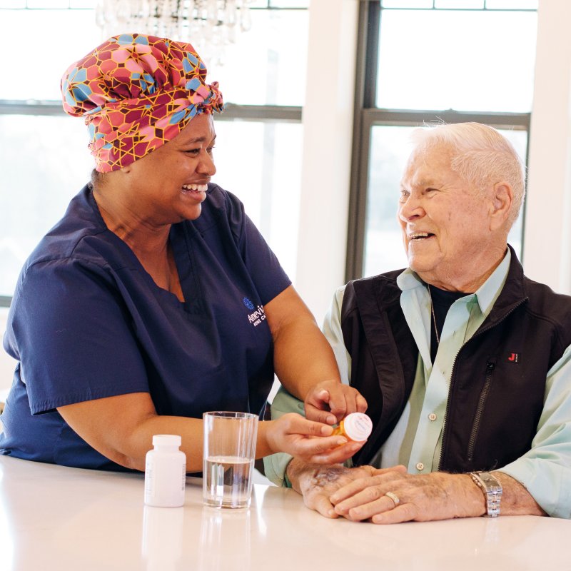A smiling female caregiver wearing an Amevian Home Care uniform points to a prescription bottle while assisting an elderly man seated at a table.