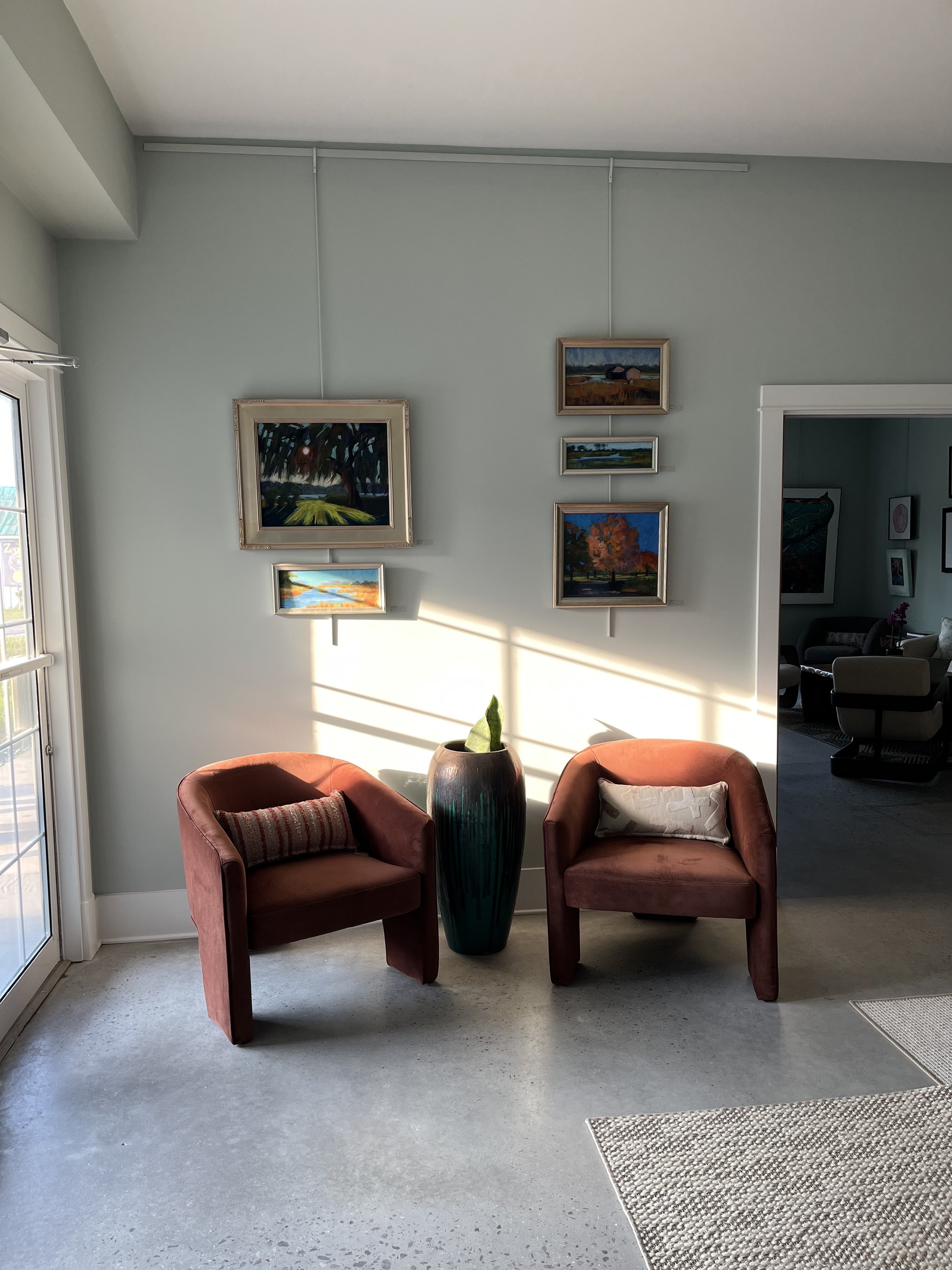 Living room corner with two terracotta armchairs, a tall green vase with a plant, wall art, and sunlight casting shadows.