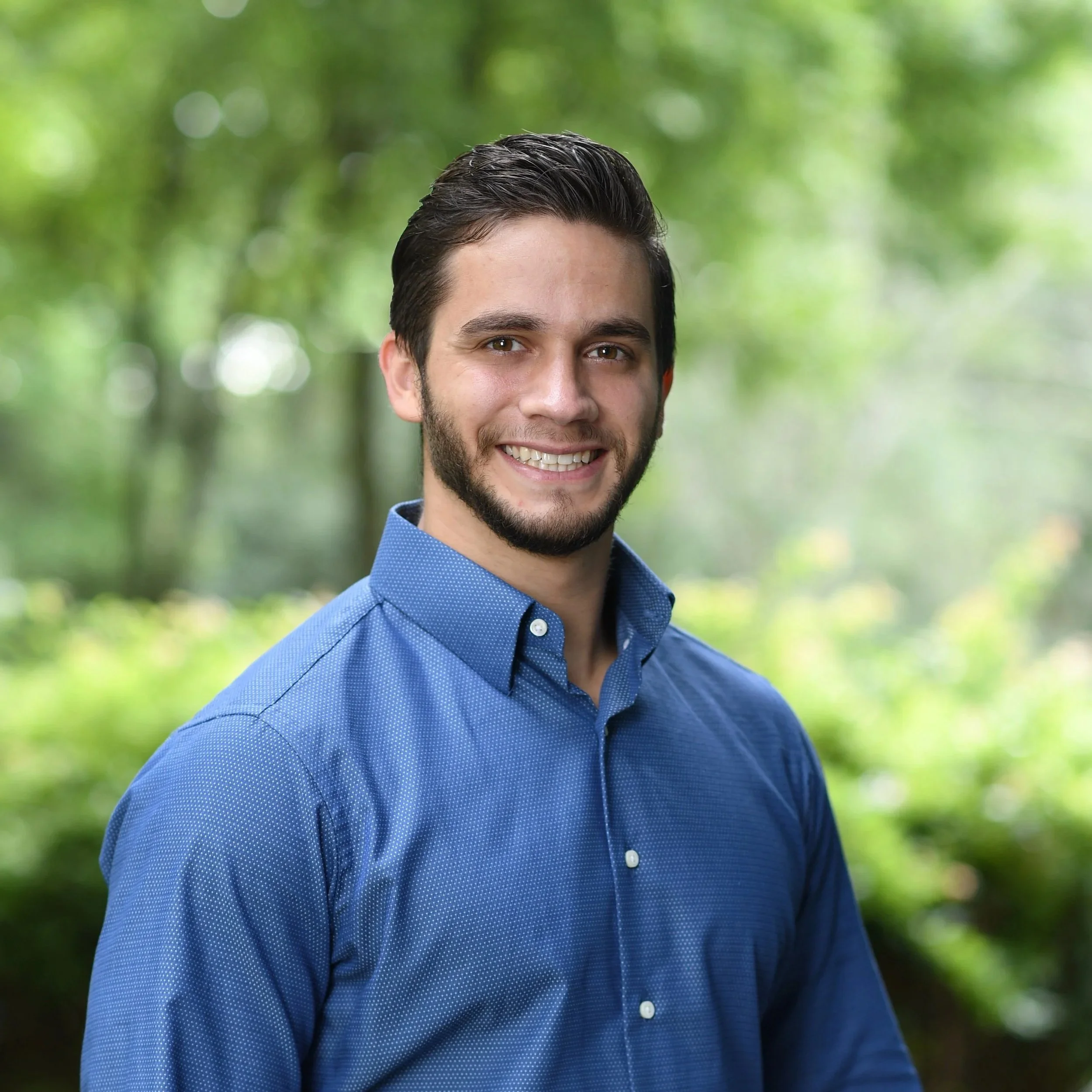 A smiling young man with dark hair and facial hair, wearing a blue button-up shirt, standing outdoors against a blurred green background of trees.
