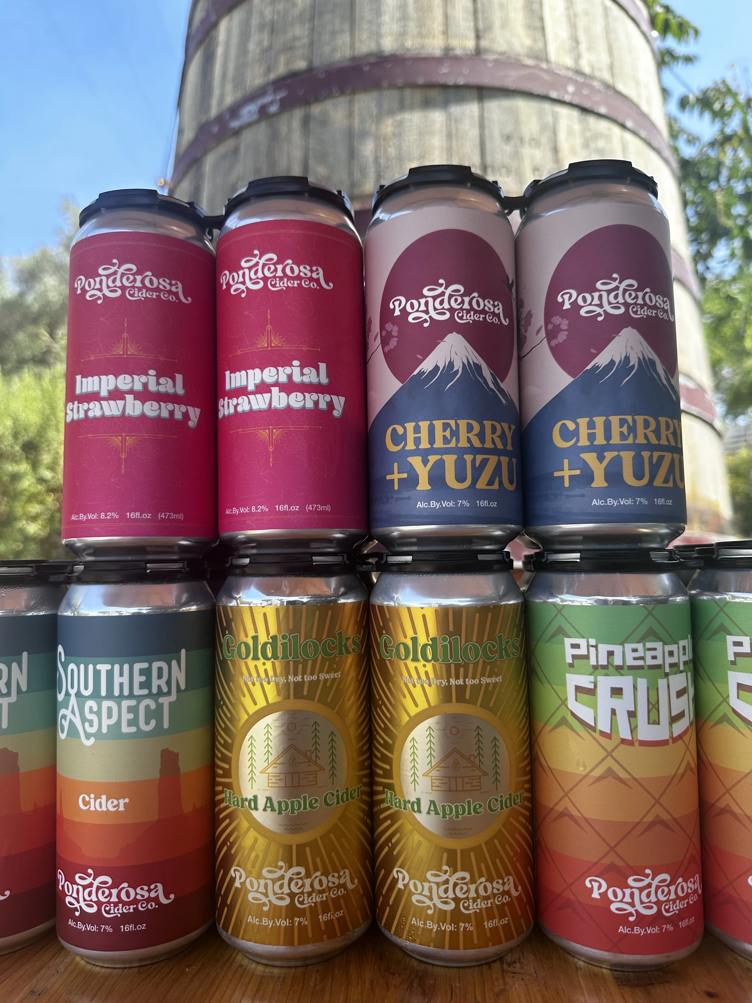 A bottle and five cans of Ponderosa Cider Co. beverages on a wooden surface outdoors. The bottle is labeled "Tamarco" and is aged on French oak. The cans include flavors like Hopped Cider, Dry Cider, Fruited Cider, Pineapple Cider, and another Dry Cider, each with colorful designs.