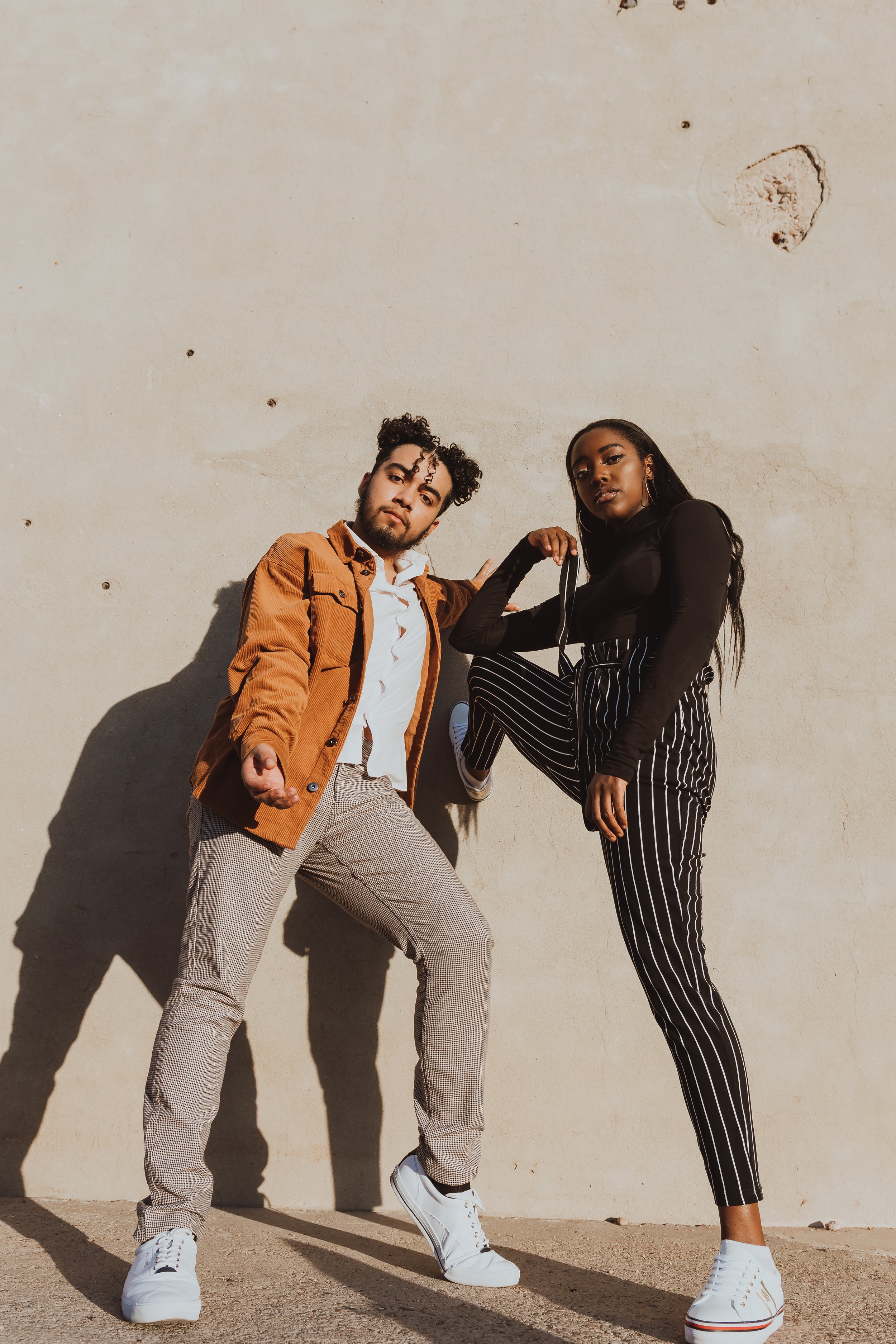 Two stylish individuals posing against a wall, one wearing a brown jacket and the other in striped pants, both with white sneakers.