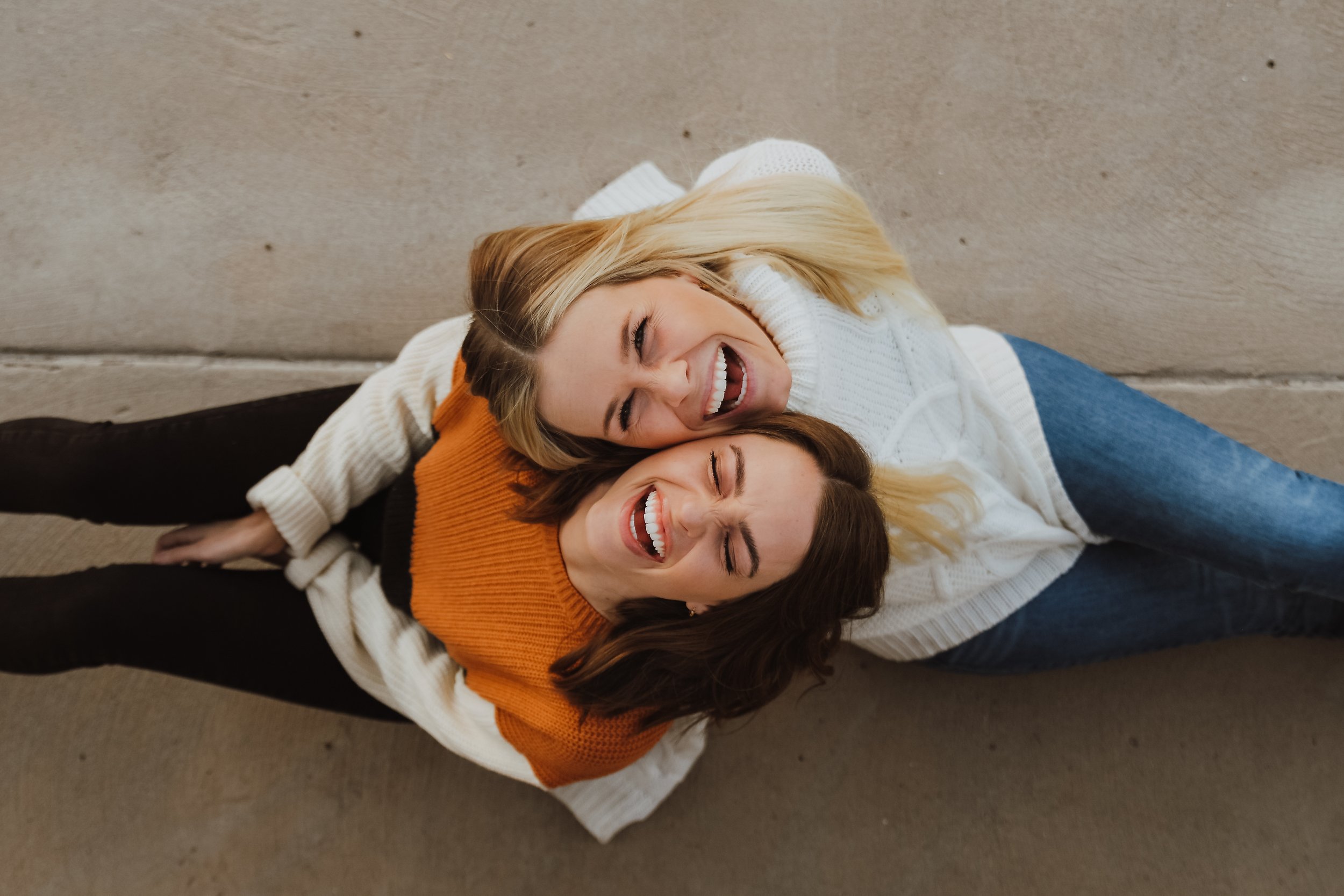 Two women laughing and embracing, viewed from above. One is wearing a white sweater and jeans, the other an orange sweater and black pants.