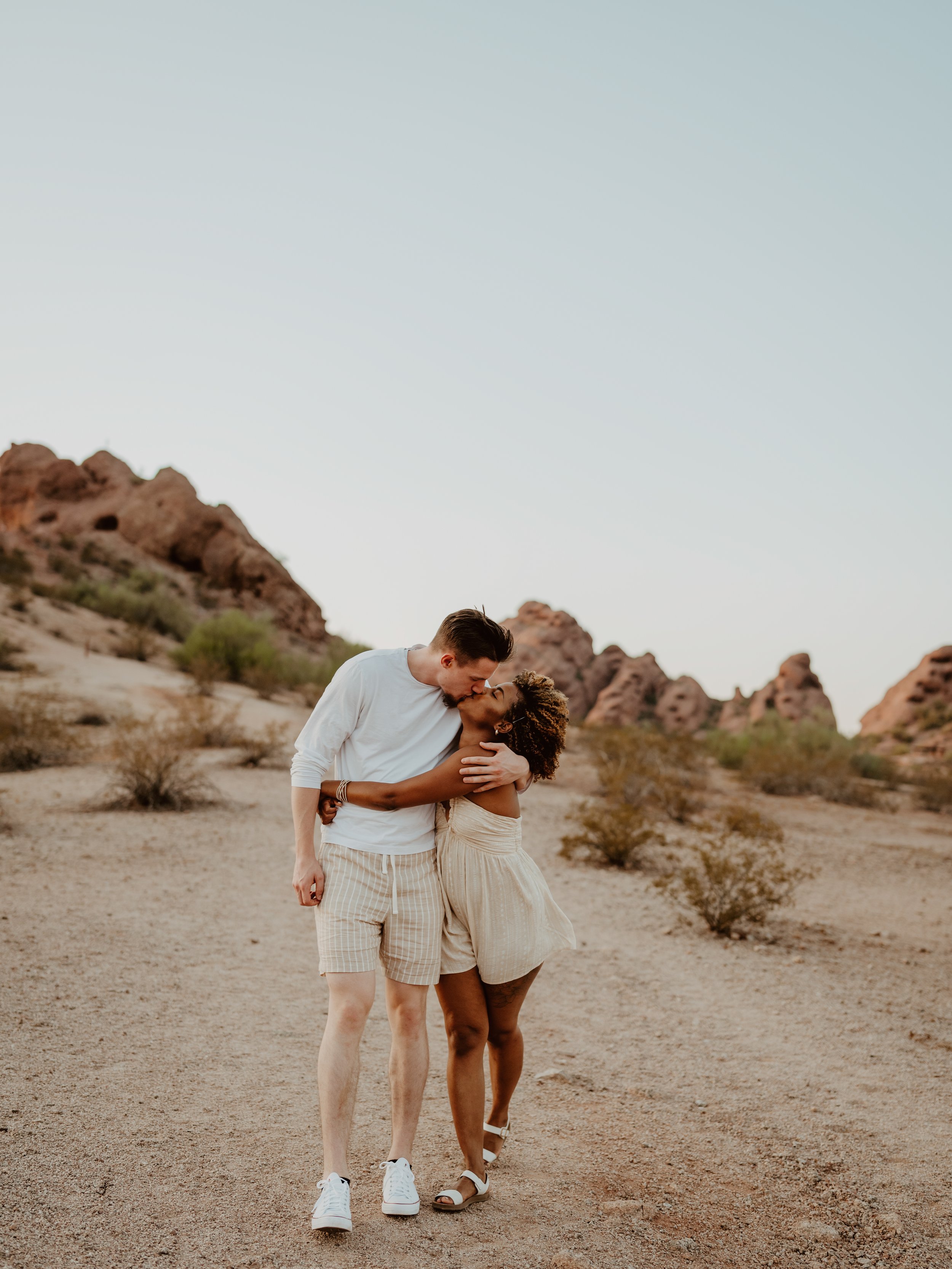 A couple embracing and kissing in a desert landscape with rocky formations in the background.