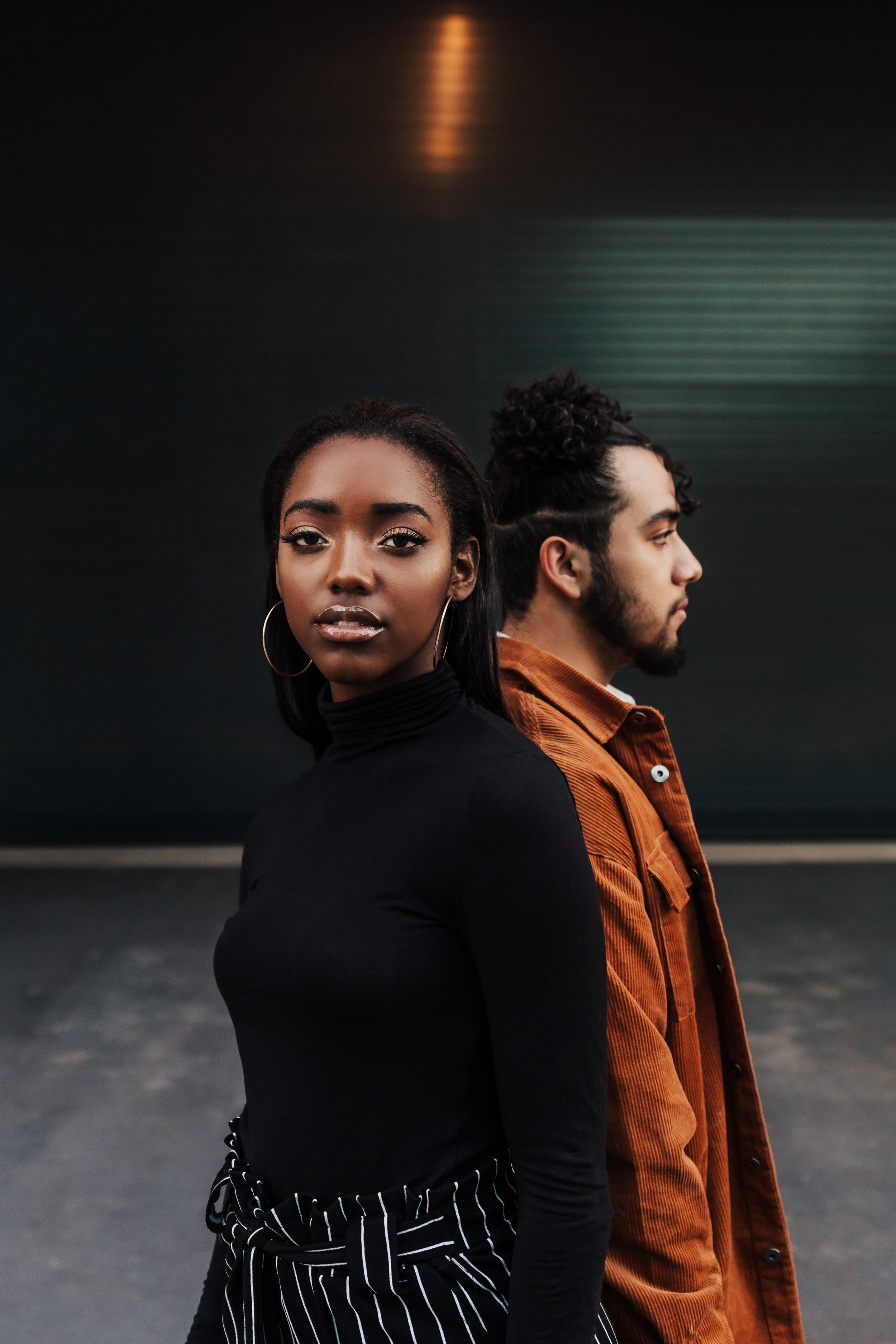 Two people standing back-to-back, one in a black turtleneck and hoop earrings, the other with a beard and wearing a brown corduroy jacket.