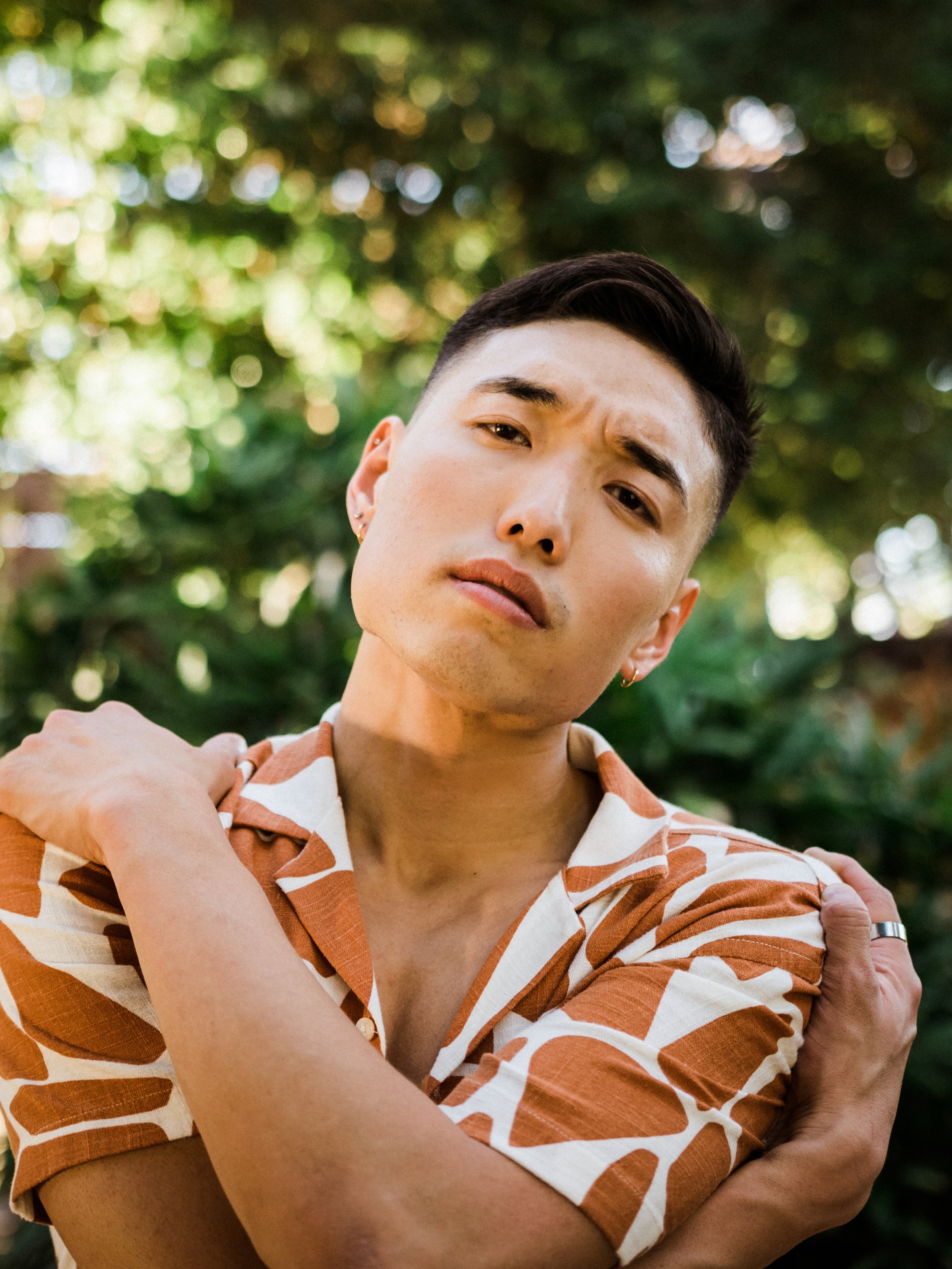 Person in patterned shirt with crossed arms and a thoughtful expression, standing outdoors with blurred greenery in the background.