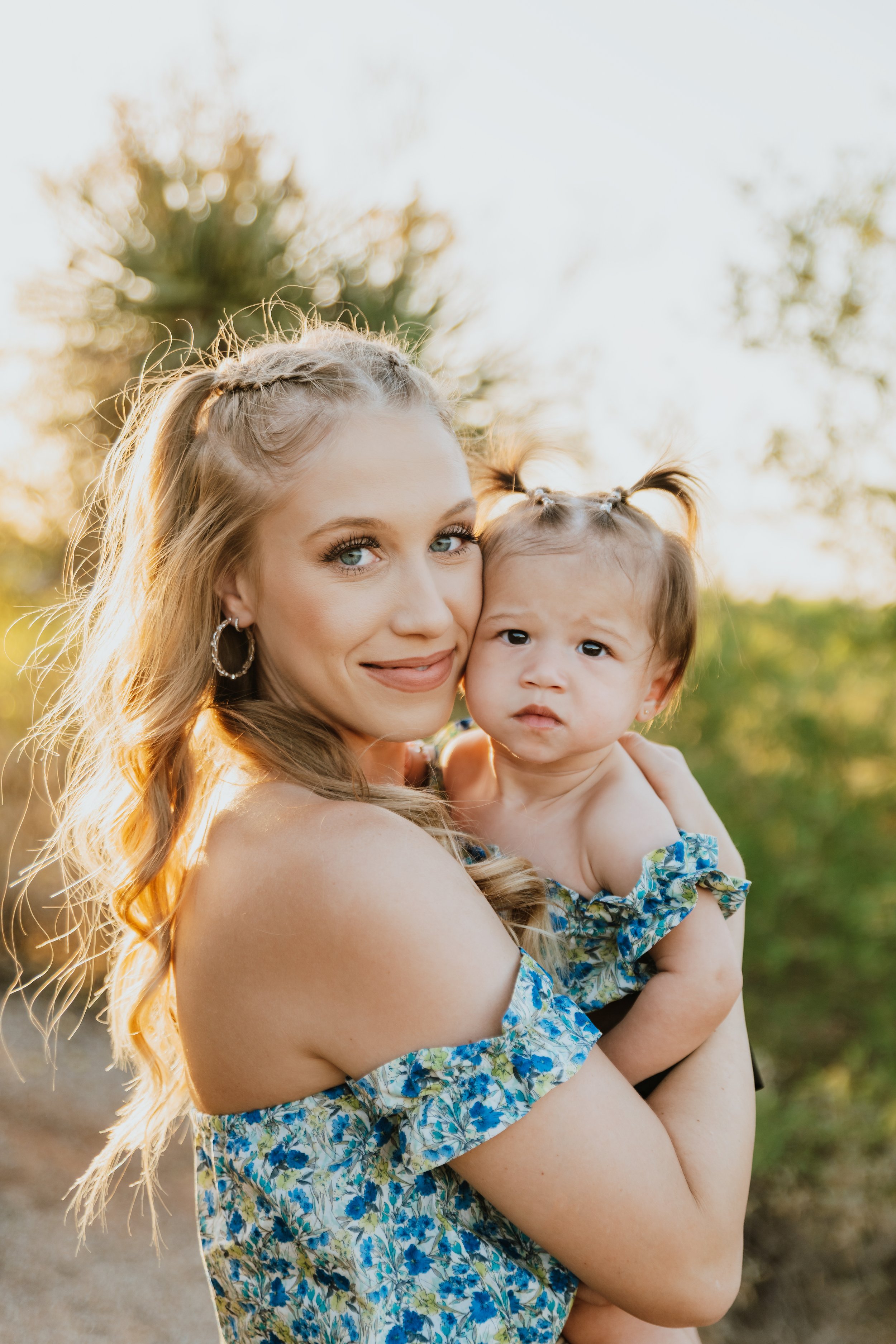 Woman holding a baby, both wearing blue floral dresses, in an outdoor setting with soft sunlight.