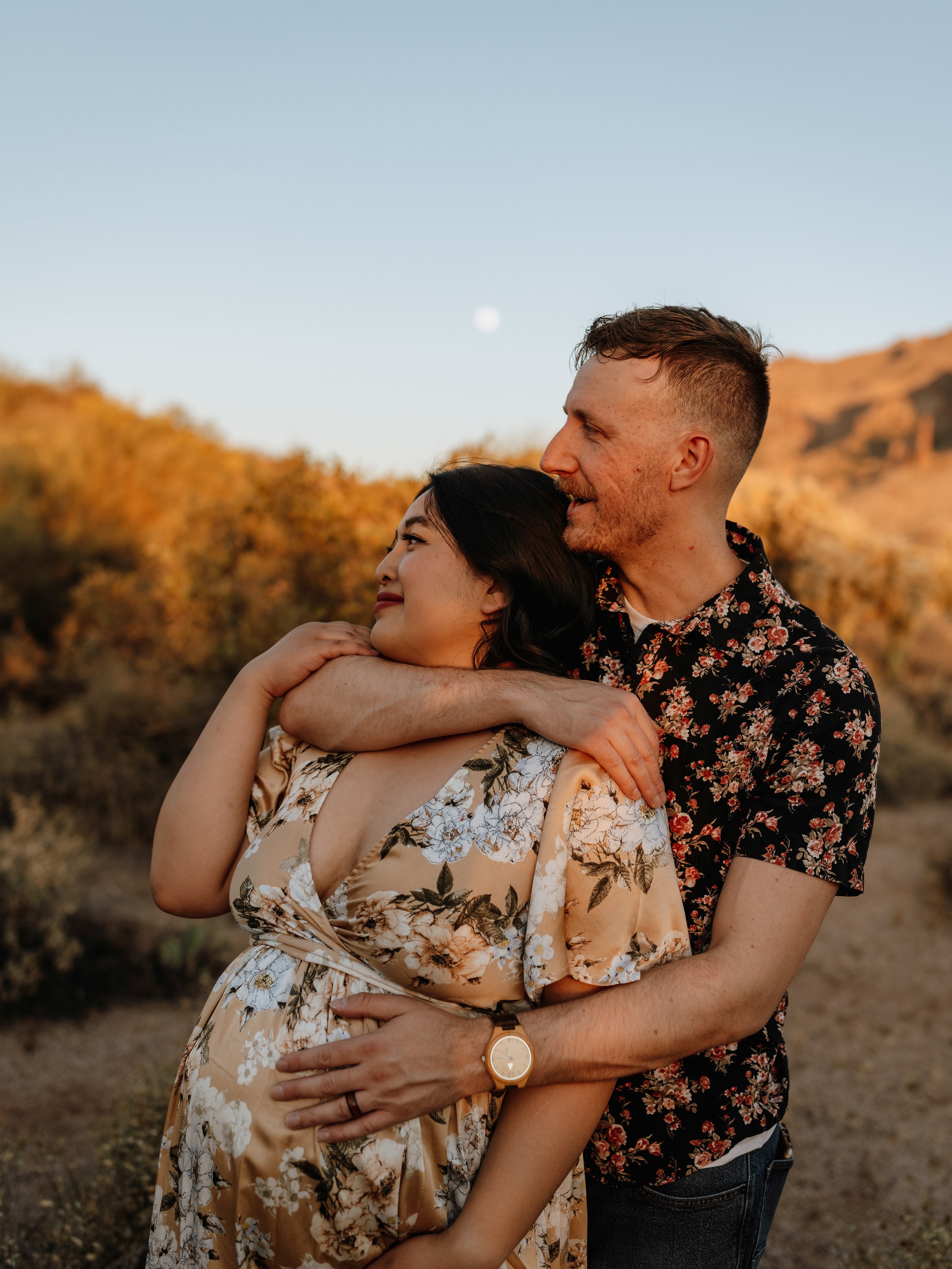 A couple embracing in a landscape with a clear sky. The woman is pregnant and wearing a floral dress, while the man is wearing a floral shirt. The background shows hills and vegetation at sunset.