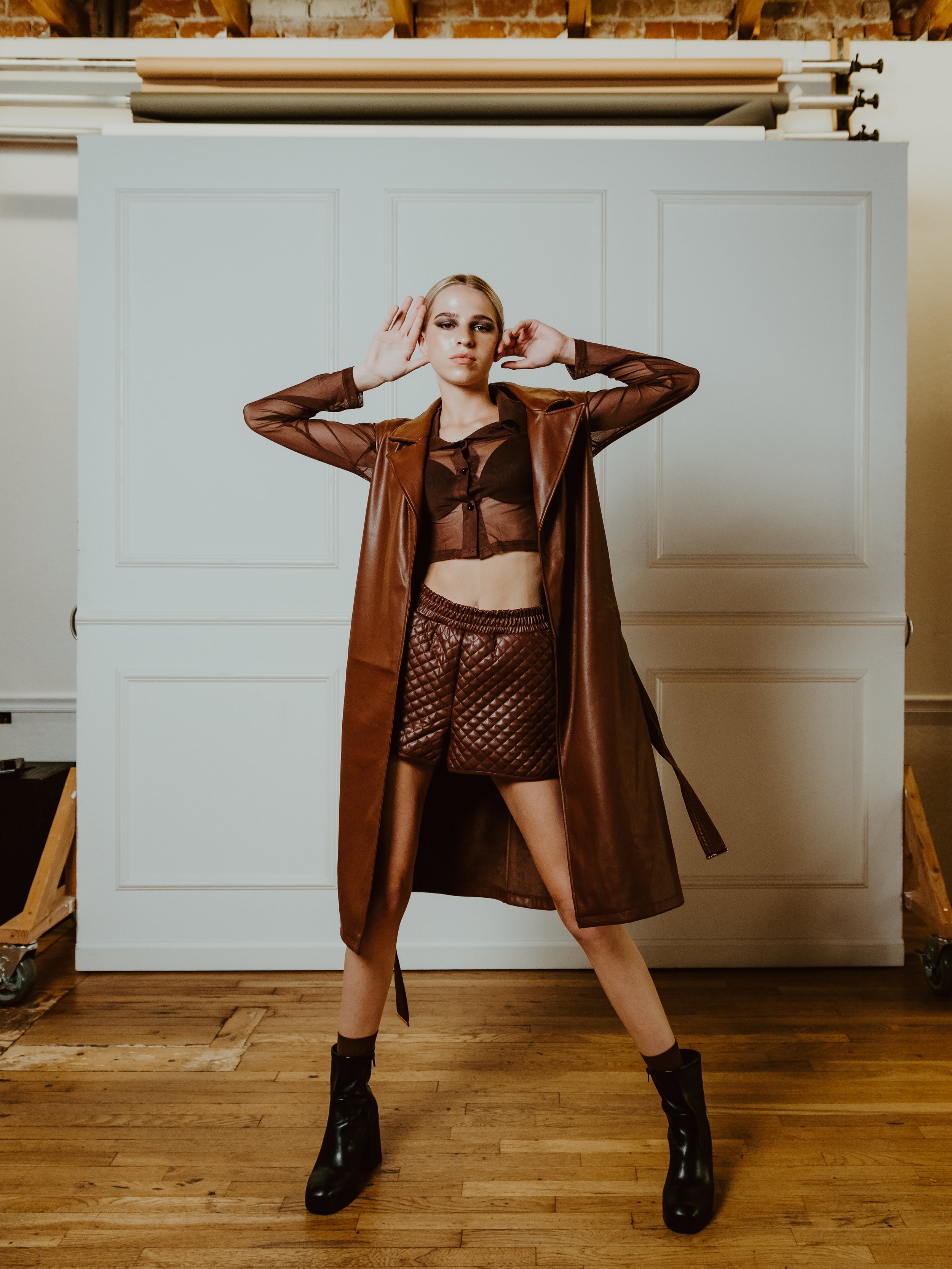 Fashion model wearing a brown leather trench coat, sheer blouse, and quilted shorts, posing indoors with hands near head.