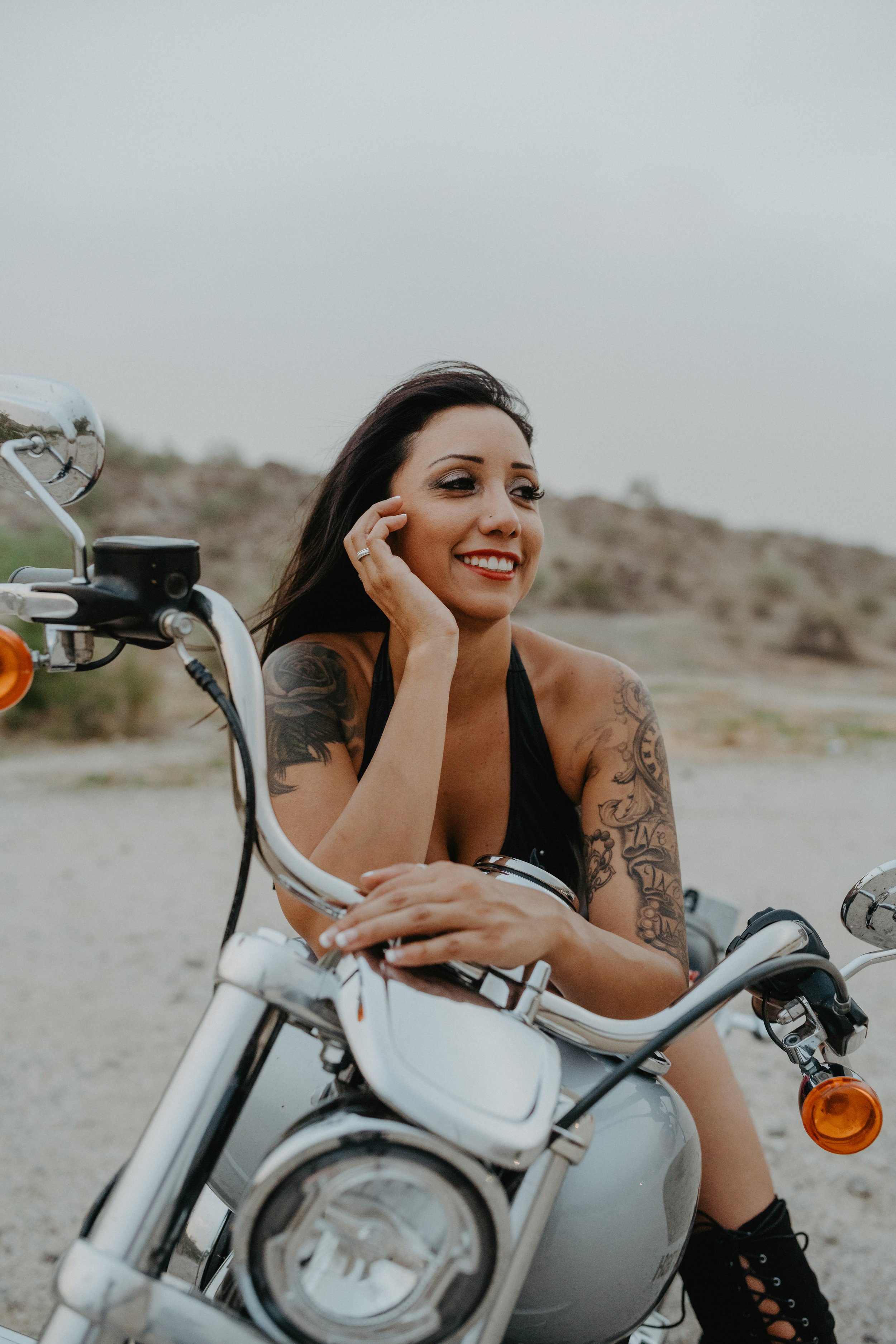 Woman with tattoos sitting on a motorcycle, smiling, outdoors.