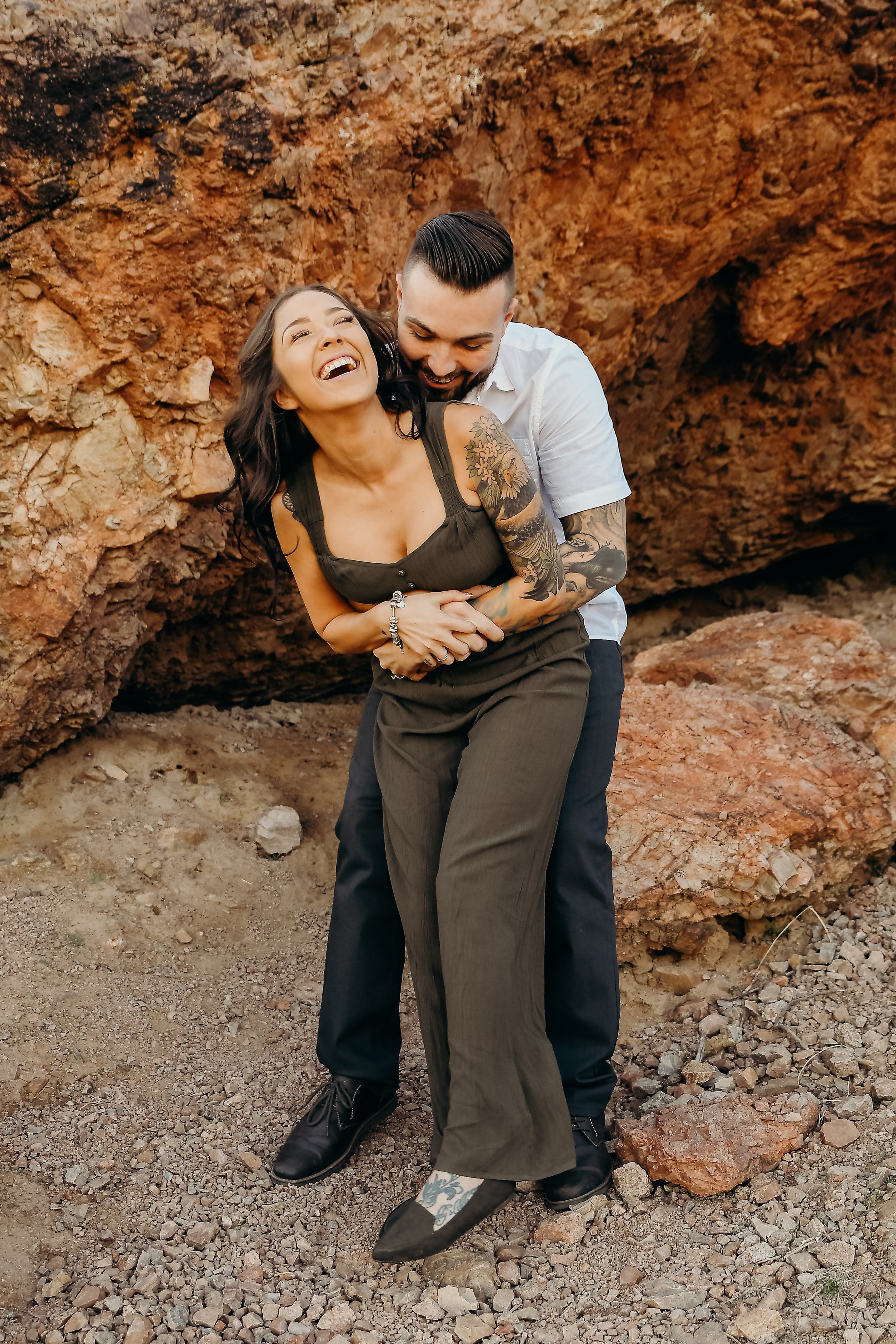 A couple embraces joyfully in front of a rocky background. The woman is wearing a dark sleeveless outfit with visible tattoos, while the man is dressed in a white shirt, holding her from behind.