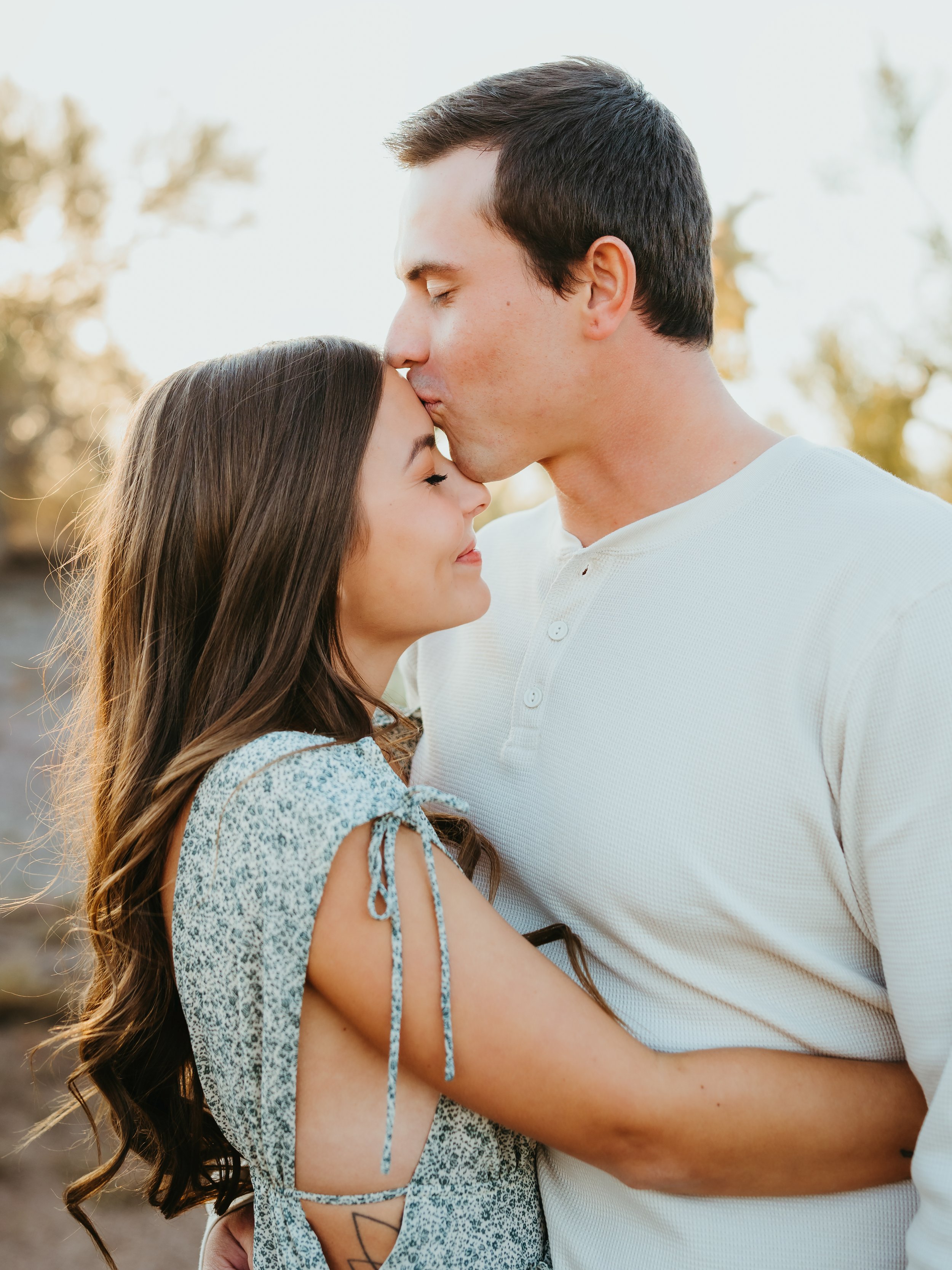 A couple embracing, with the man kissing the woman's forehead. The woman wears a floral dress, and the man wears a white shirt. They are in an outdoor setting with trees in the background.