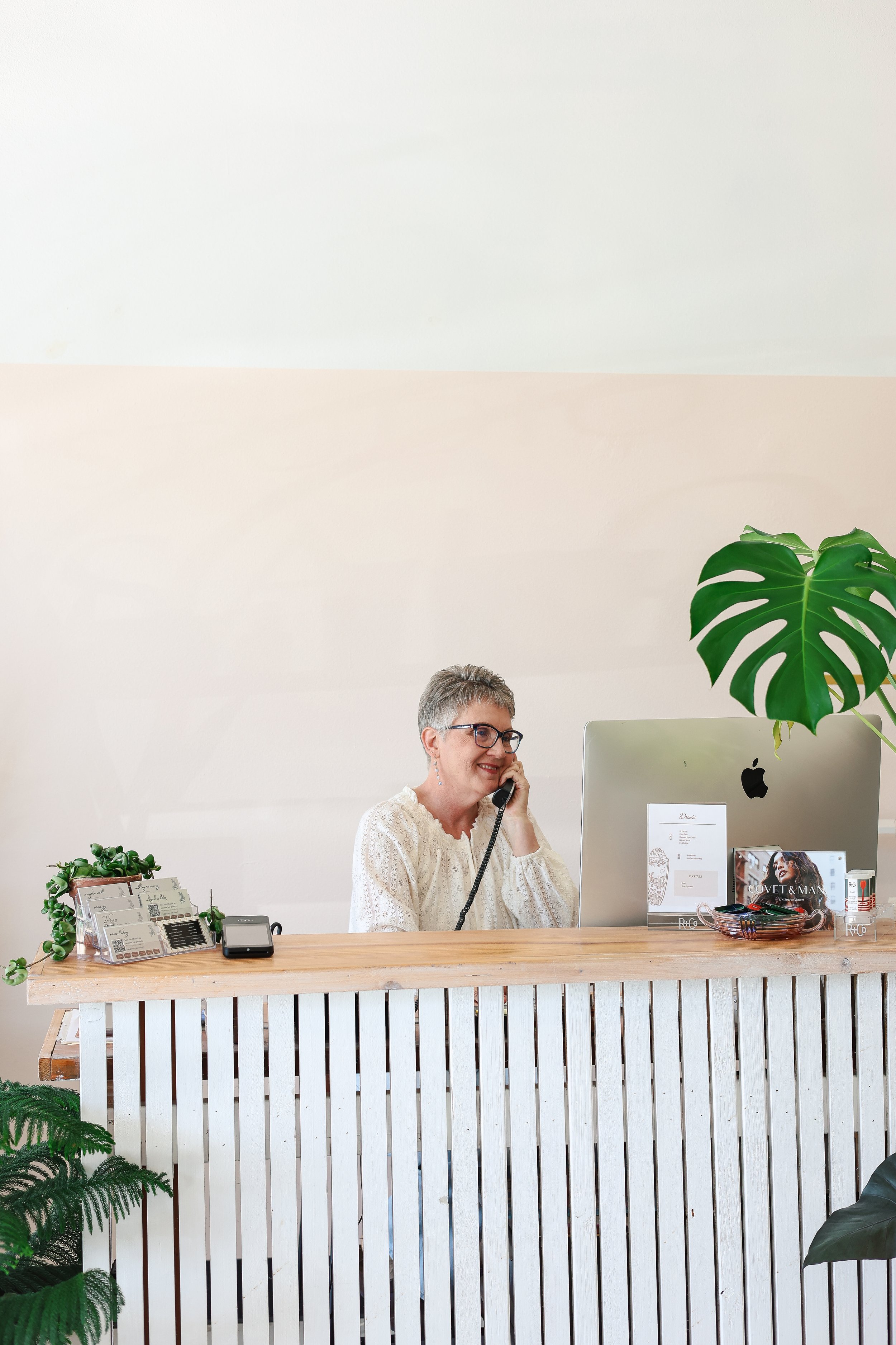 A smiling receptionist with short grey hair talking on the phone at a white slatted wooden front desk in a bright, modern office.