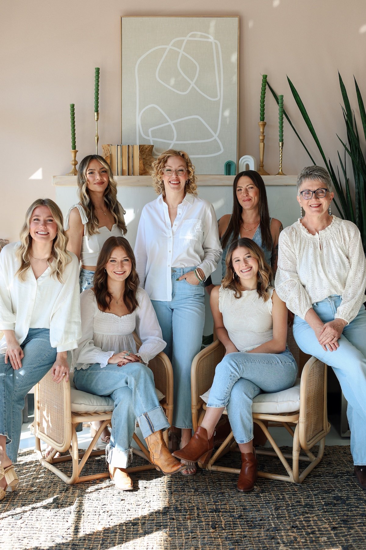 Seven smiling women of the Dwell salon team posing together in coordinated white tops and light blue jeans in a bright, stylish salon interior.