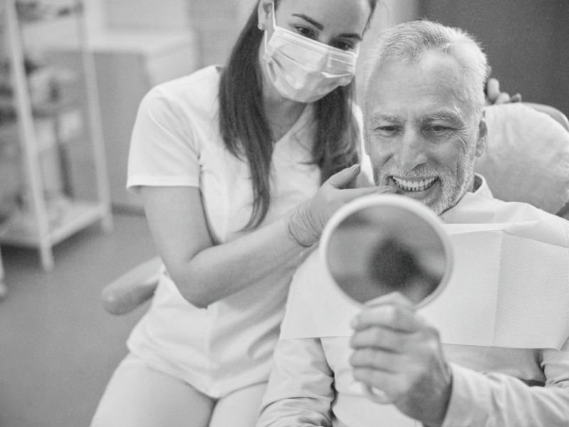 An old man admiring his new dental implants while looking in a handheld mirror while a dental hygenist from Northstate Dental explains how they work to him