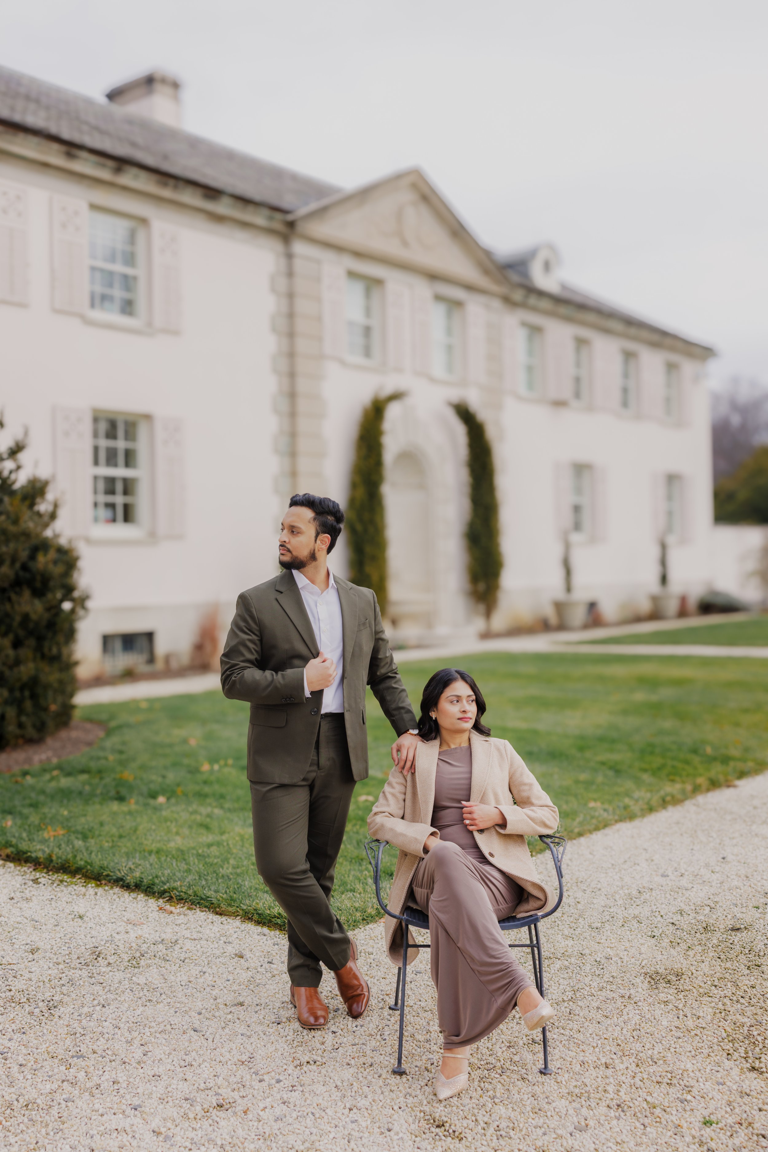 A man in a suit standing next to a woman in a dress sitting in a chair outdoors, with a large white house in the background.