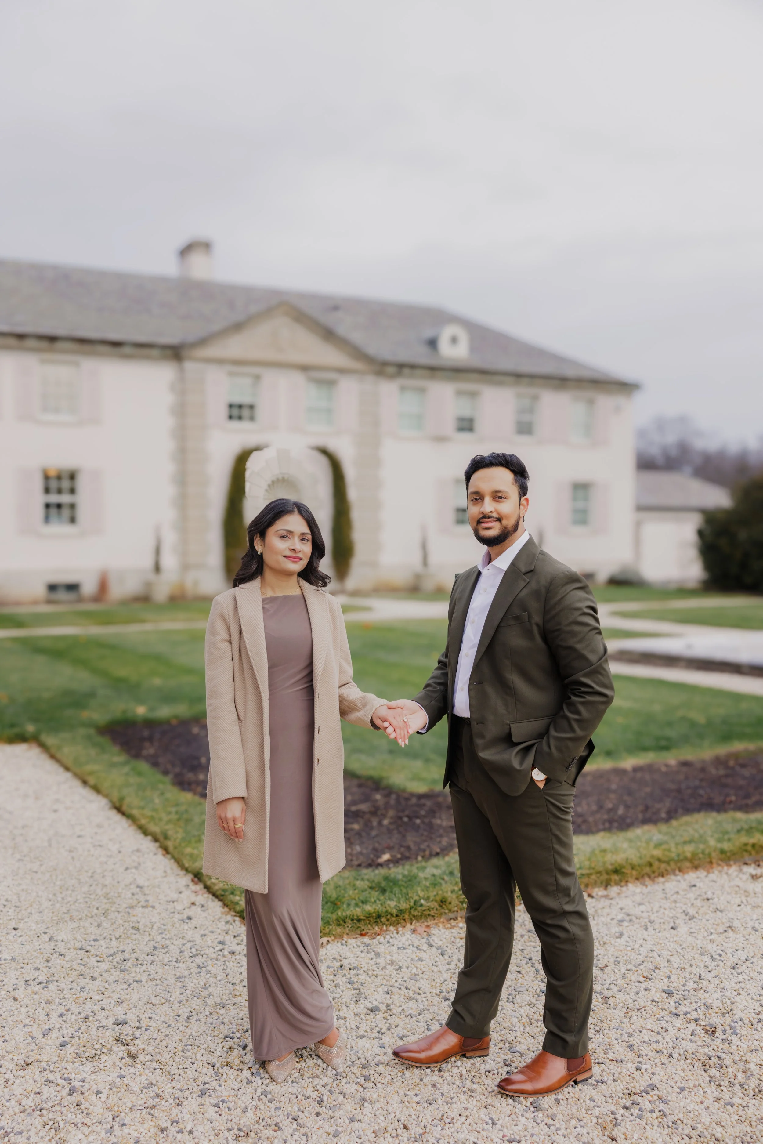 A man and woman shaking hands outside a large, elegant house with a well-maintained lawn on a cloudy day.