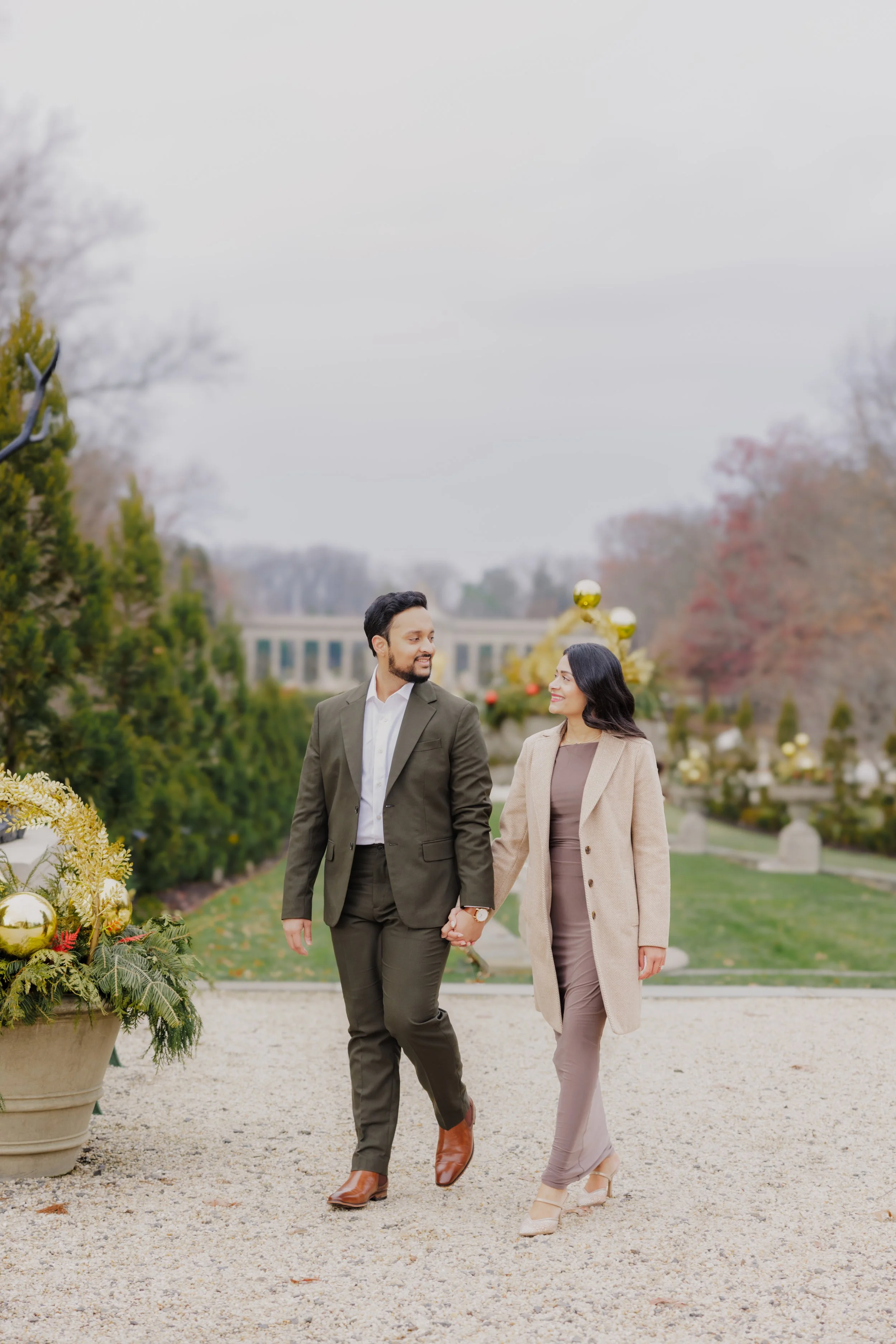 A couple holding hands and walking outdoors on a cloudy day, decorated with Christmas ornaments and greenery, with a large garden and trees in the background.