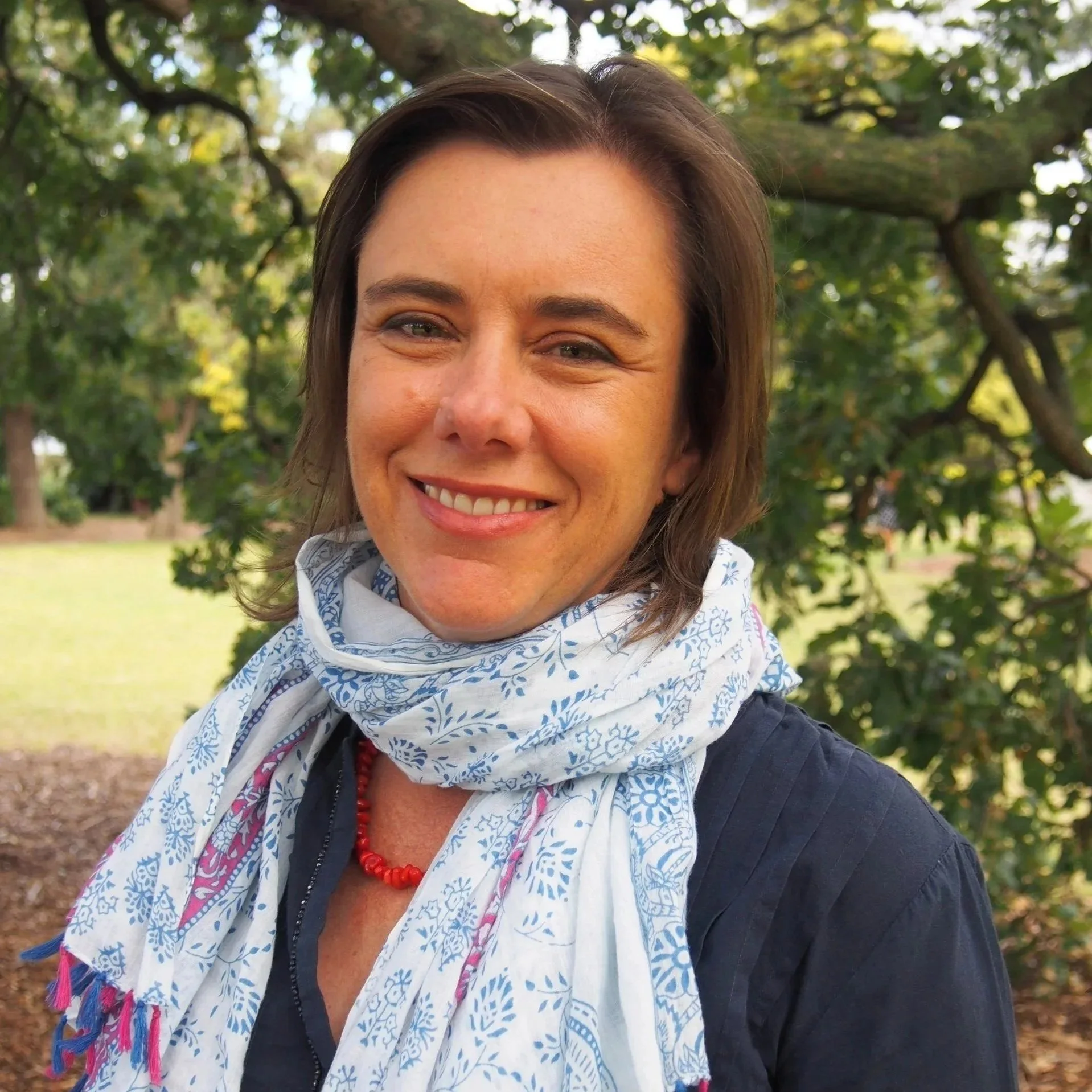 A woman taking a selfie outdoors, smiling, with green trees and sunlight in the background. She has shoulder-length brown hair and wears a white bandana with blue patterns around her neck, a dark jacket, and a red bead necklace.