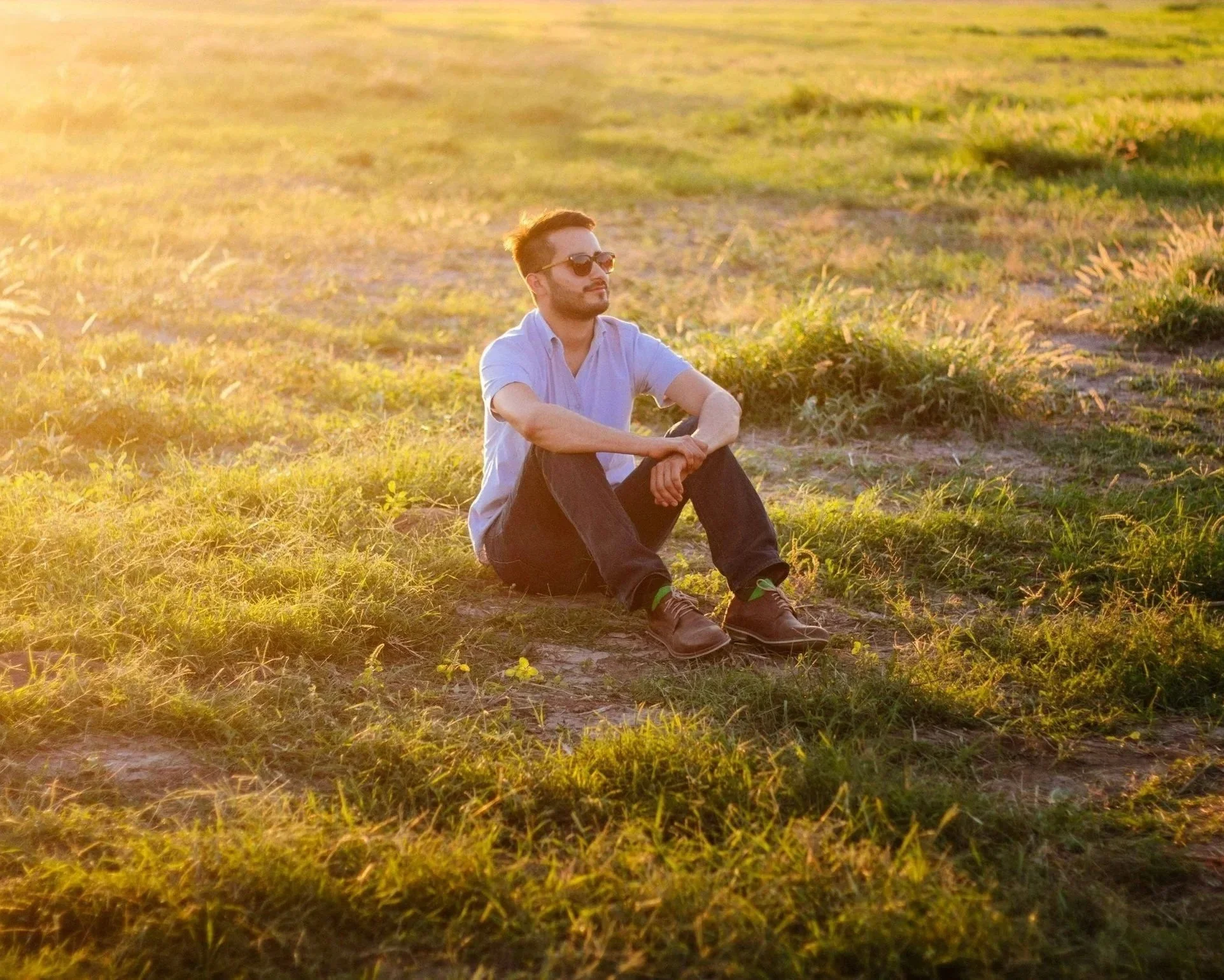A man with sunglasses, beard, and short hair sitting on grass in a field during sunset.
