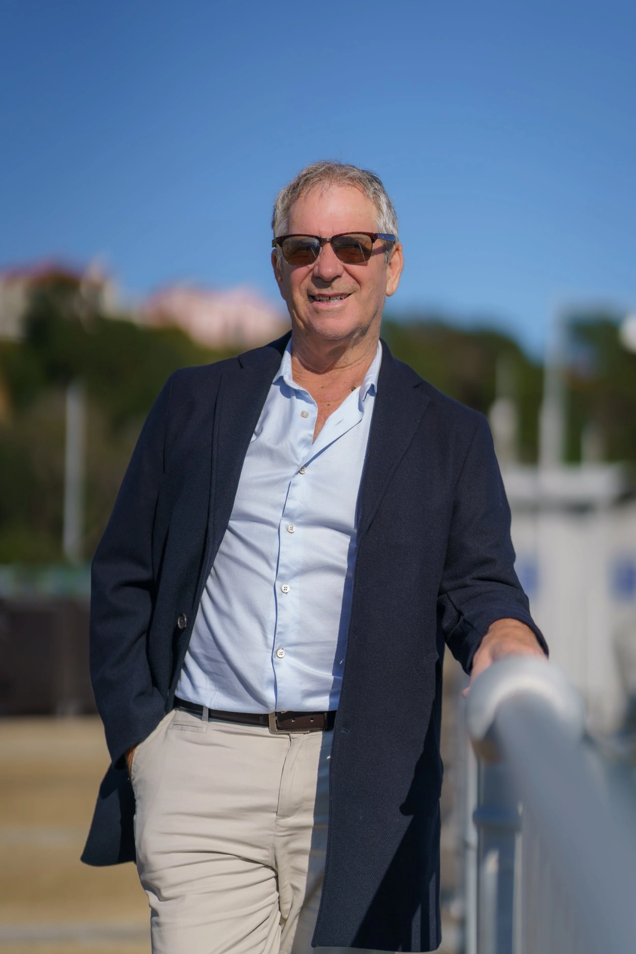 A photograph of Mike in a white button-up shirt, a dark blazer, and light beige pants, standing outdoors on a clear day with a blue sky and greenery in the blurred background.