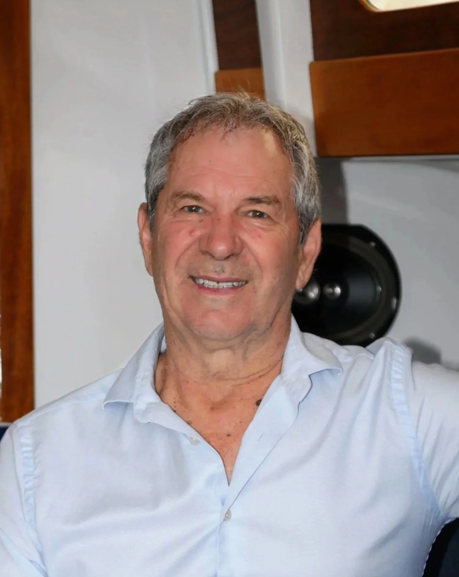 Close-up of a Mike, wearing a light blue collared shirt, sitting in a boat with a speaker and wooden furniture in the background.