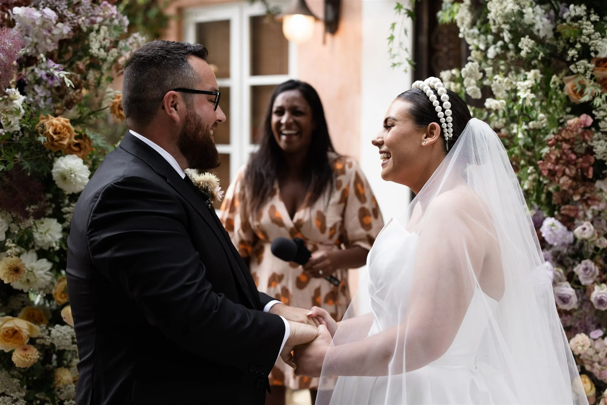 A bride and groom holding hands and smiling at each other during a wedding ceremony, with an officiant standing behind them, surrounded by floral decorations.
