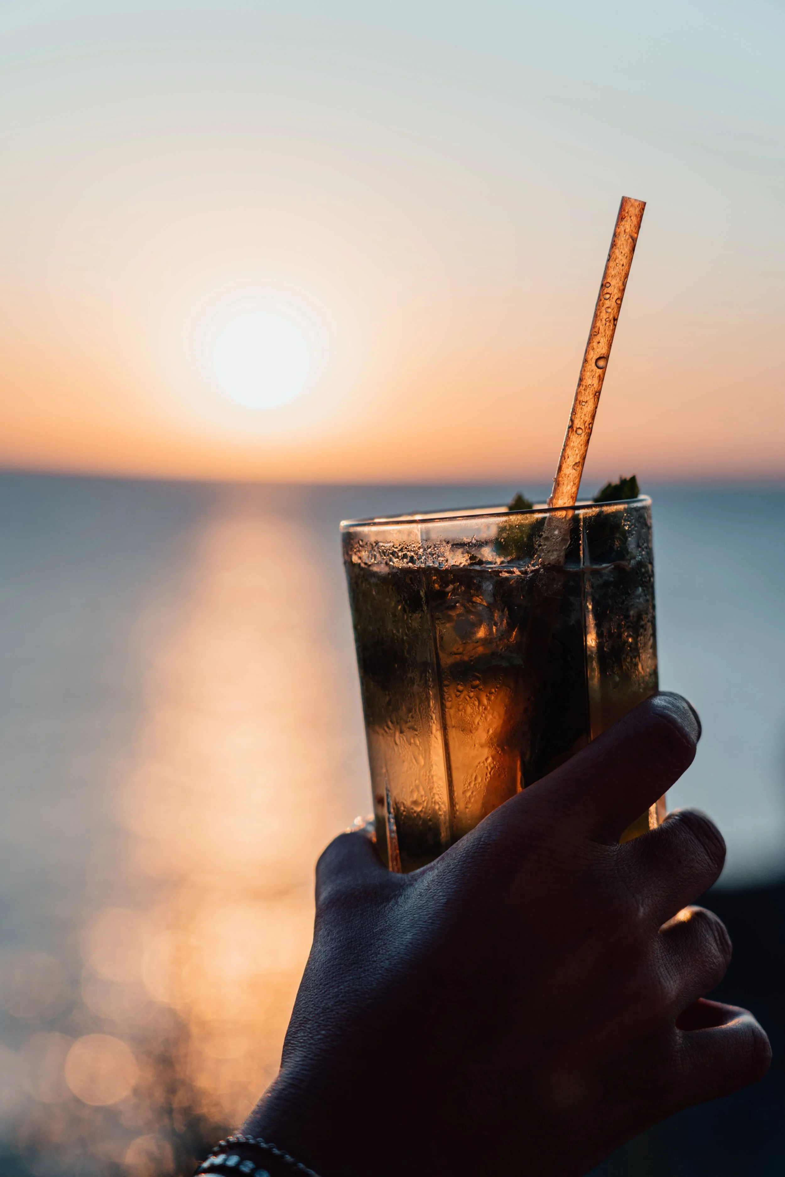 Una mano sosteniendo un vaso con bebida y hielo, con una pajilla, frente a un atardecer sobre el mar.