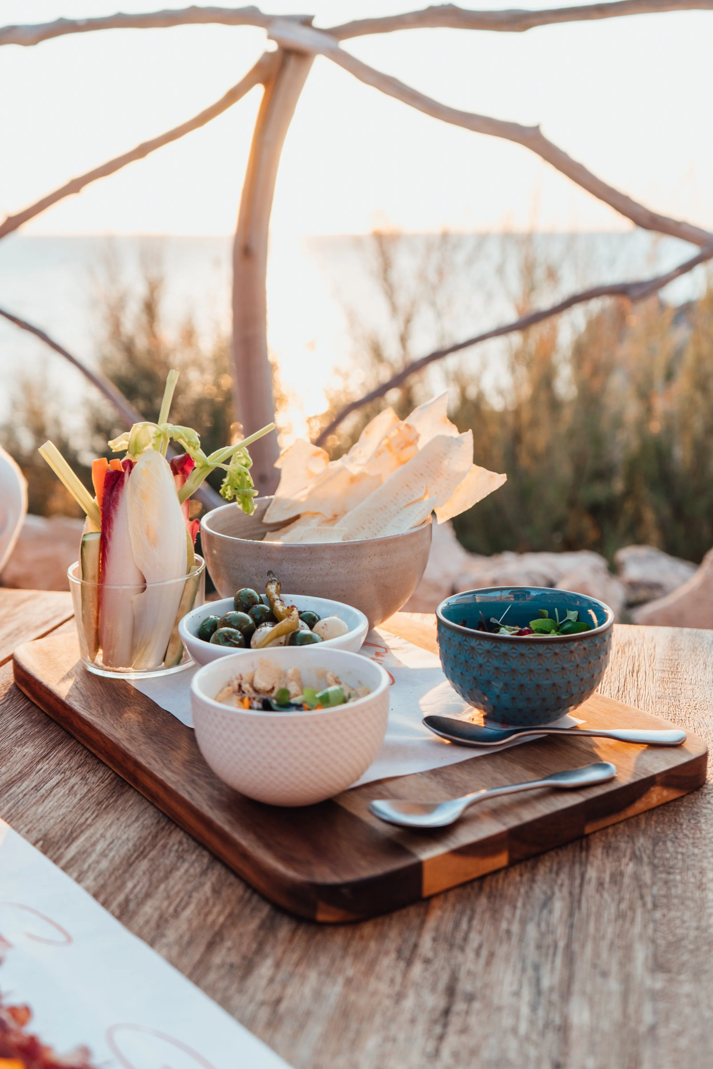 Una bandeja de madera con varios platos pequeños con ensalada, frutas, y chips, en un ambiente exterior durante el atardecer.