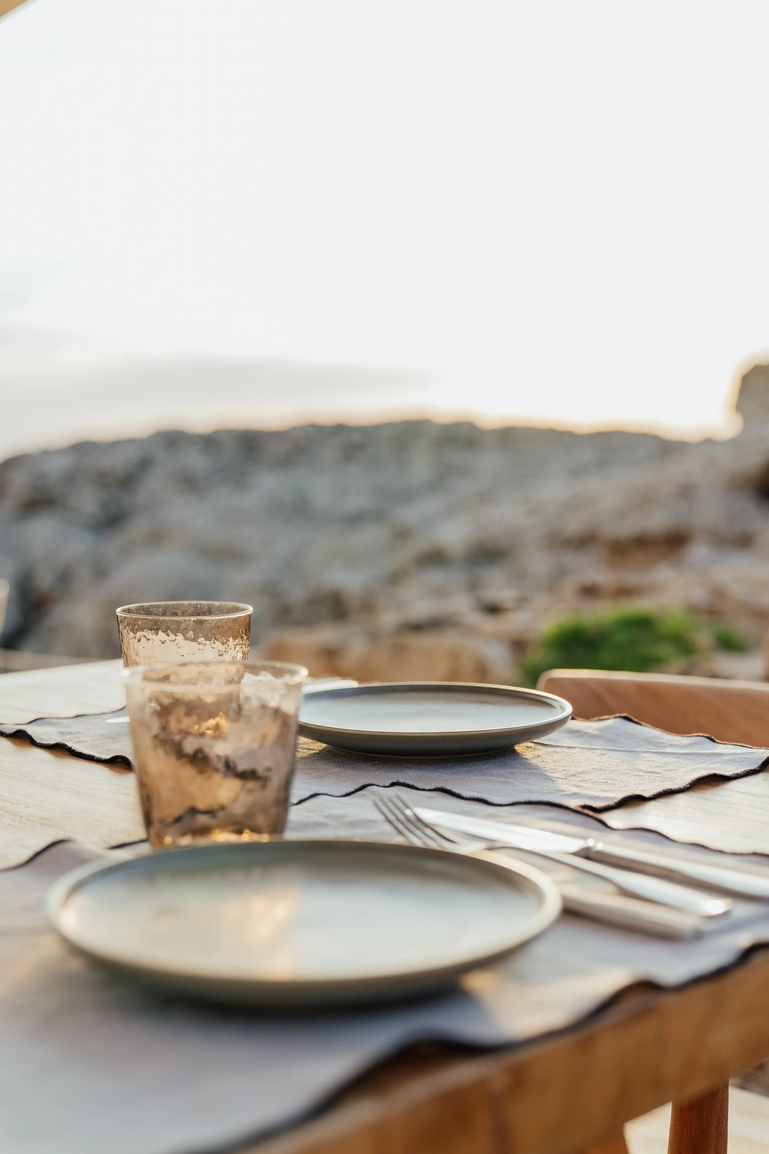 Mesa de comedor al aire libre con platos, vasos y cubiertos, en un escenario natural con rocas y vegetación al fondo, durante el atardecer.