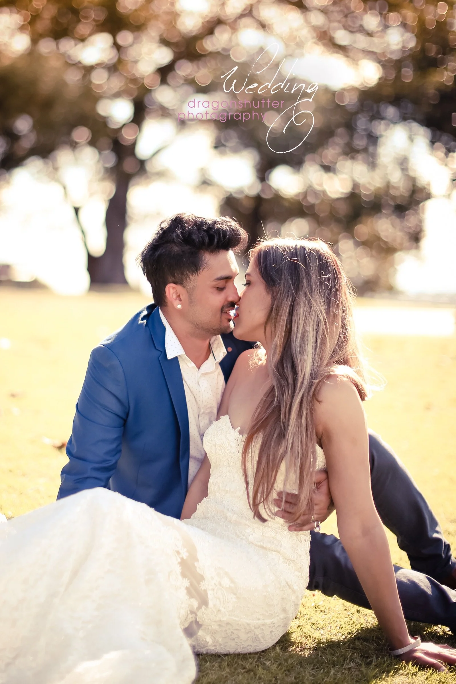 A couple sitting on the grass outdoors, about to kiss, with a blurred background of trees. The woman is in a white lace wedding dress, and the man is in a blue blazer. Text on the image reads 'Wedding' and 'dragonsshutter photography'.