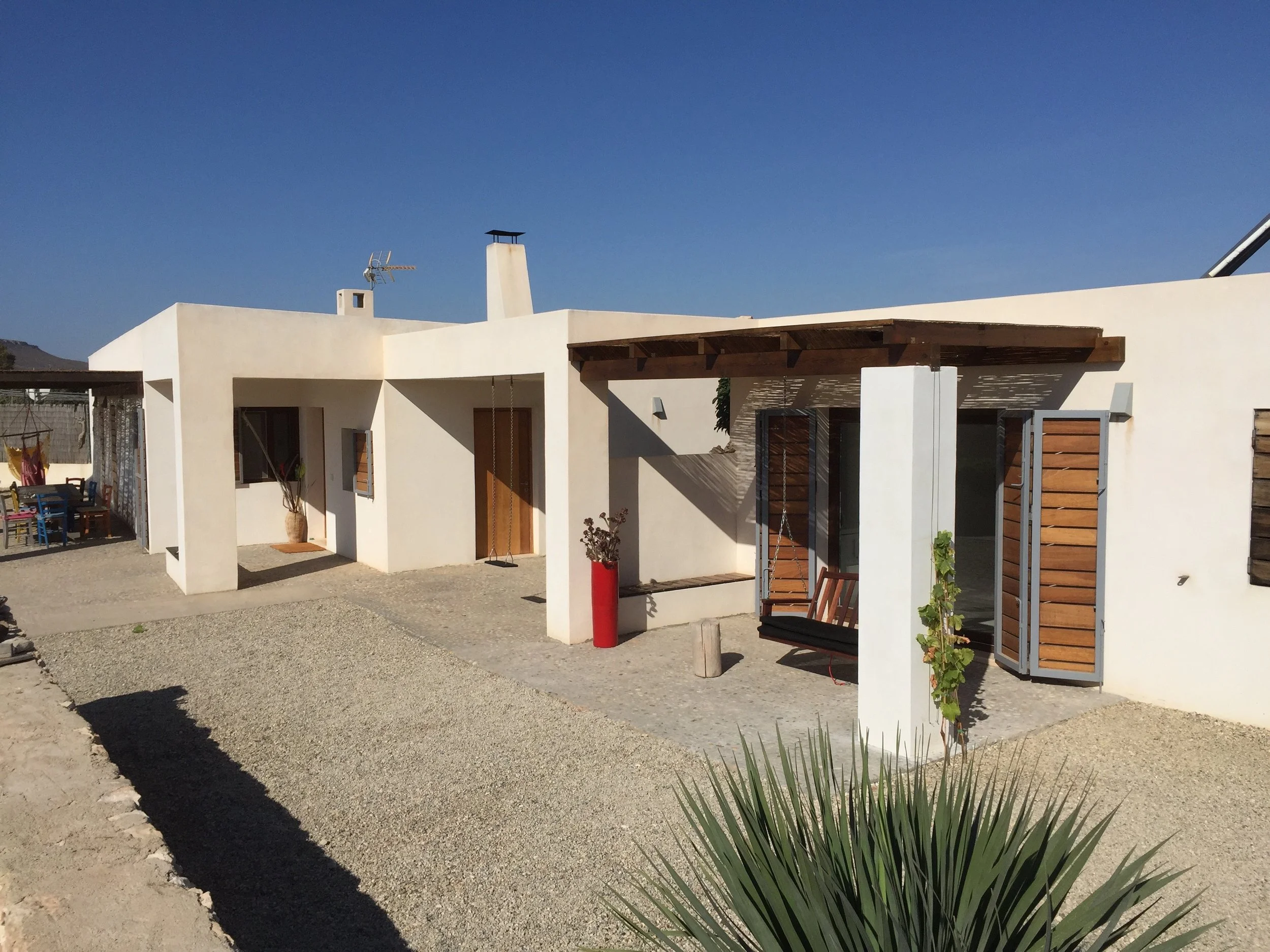 Modern white stucco house with wooden shutters, gravel yard, and a small patio area. The house has a flat roof, a chimney, and an outdoor swing. A table with chairs is visible on the patio, and a large plant is in the foreground.