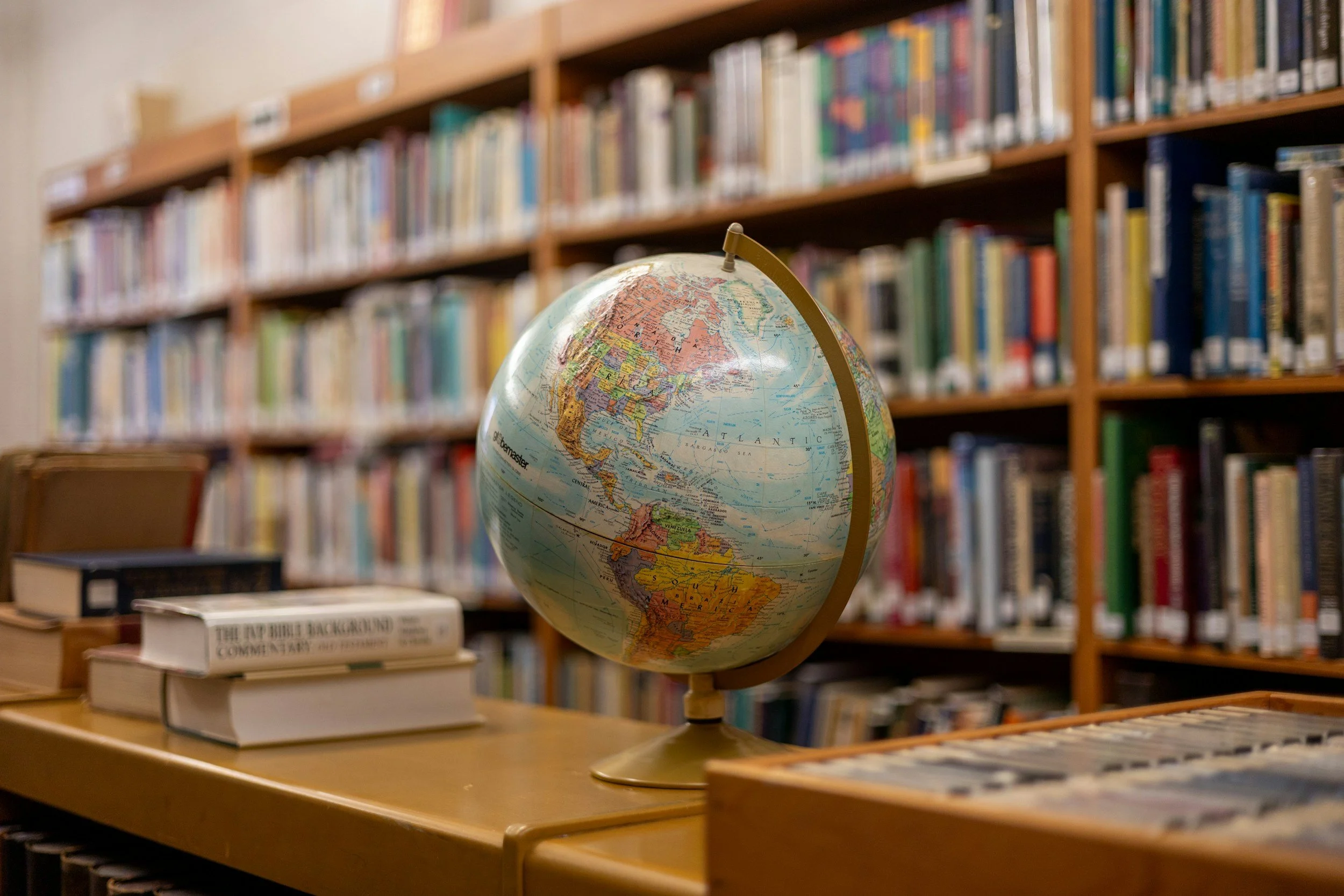 A globe on a wooden table in a library, with shelves of books in the background.
