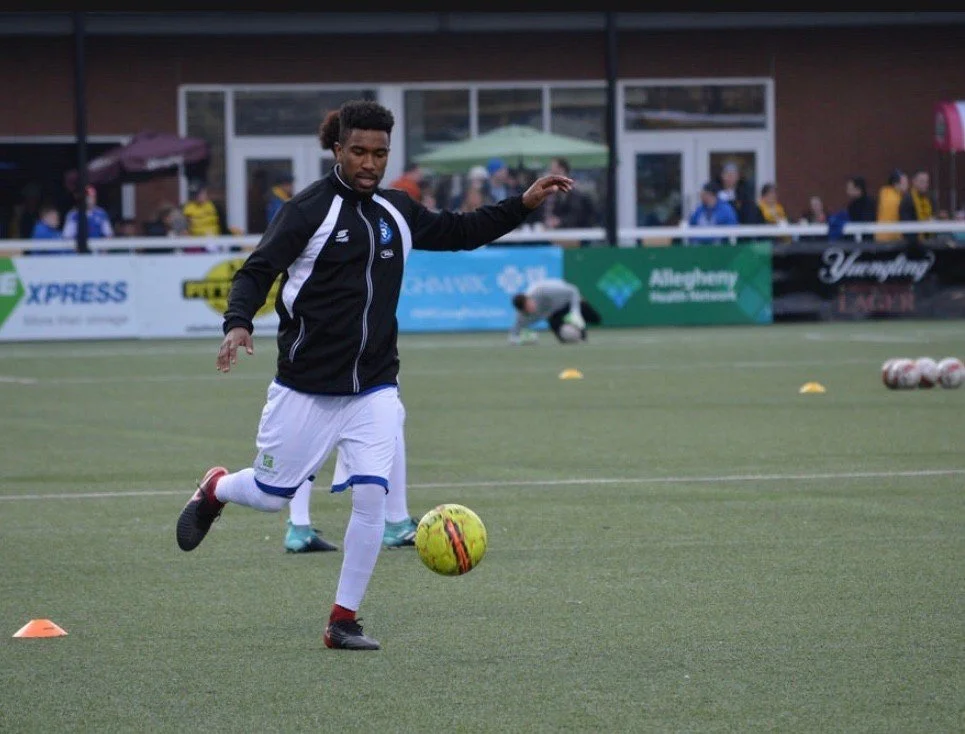 A male soccer player practicing on the field with a yellow soccer ball, wearing a black and white uniform, and there are other players and spectators in the background.