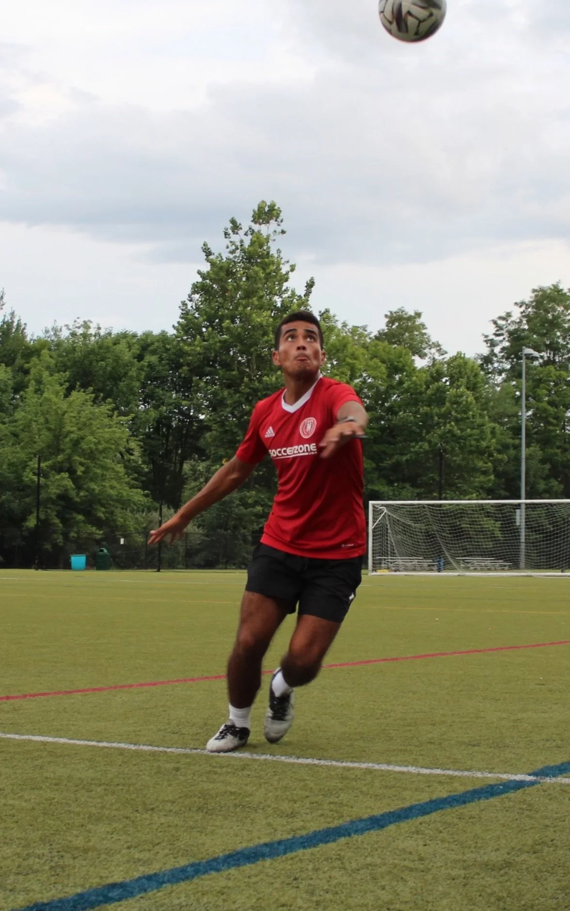A young male soccer player in a red jersey and black shorts is on a soccer field, preparing to head a soccer ball in midair, with trees and a goal in the background.